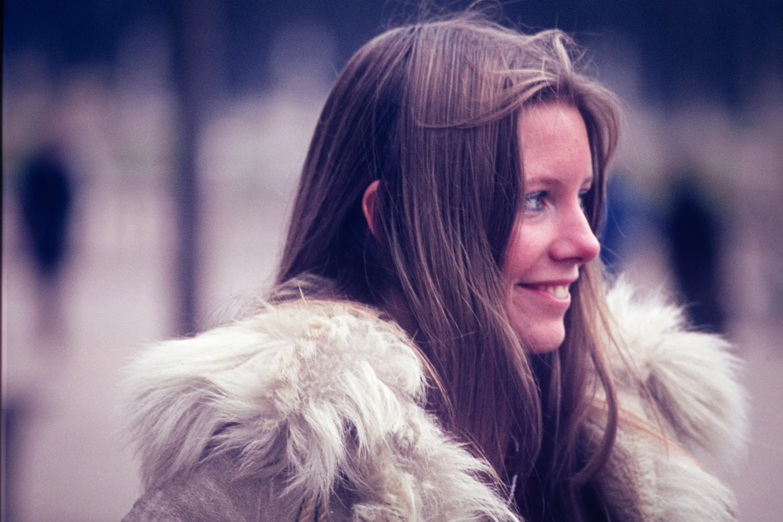 A woman with long brown hair and a smile, wearing a cream-colored fur-lined coat, looking to her right, outdoors with a blurred background.