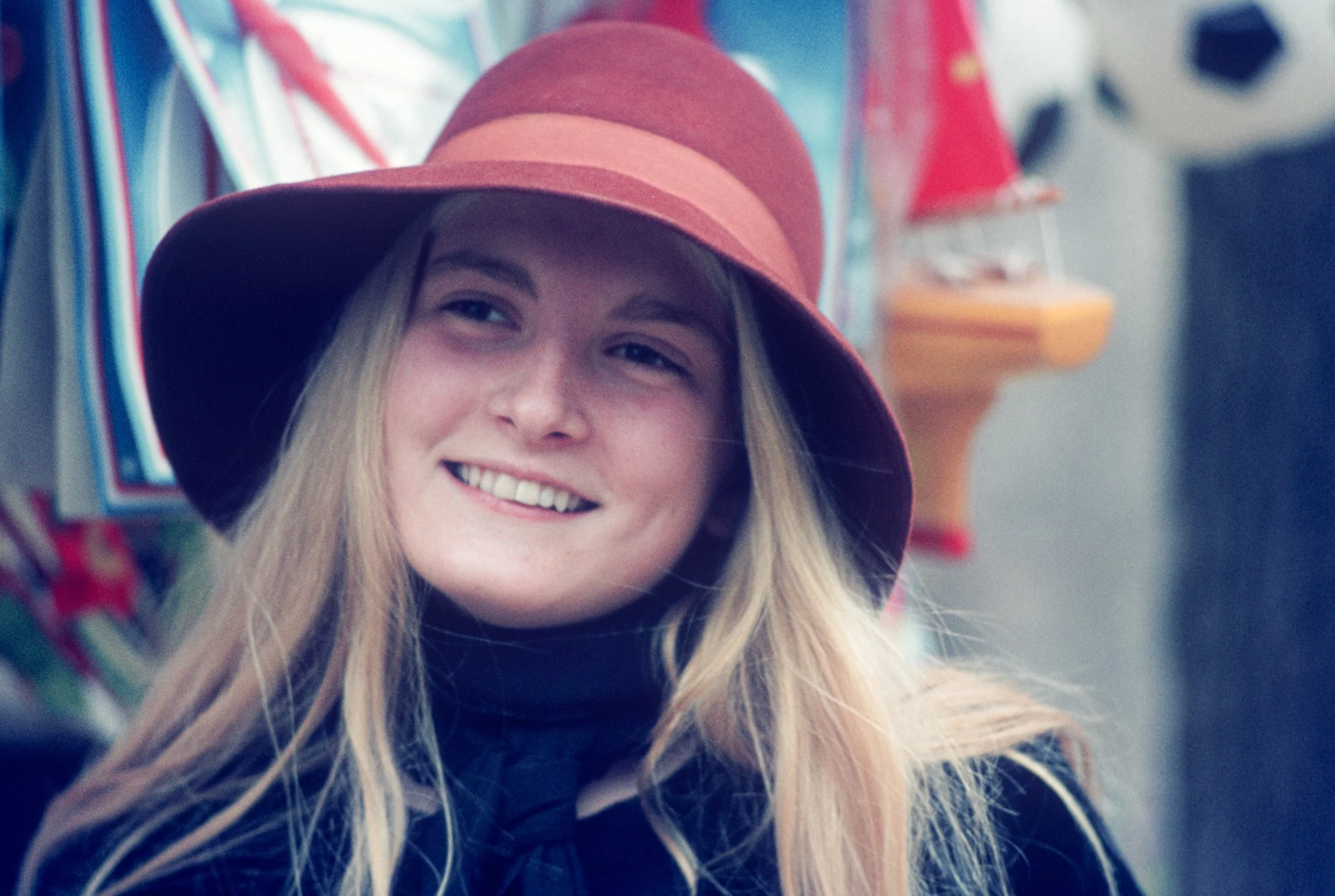 A young woman smiling wears a red wide-brimmed hat and a black turtleneck, with colorful items in the background.
