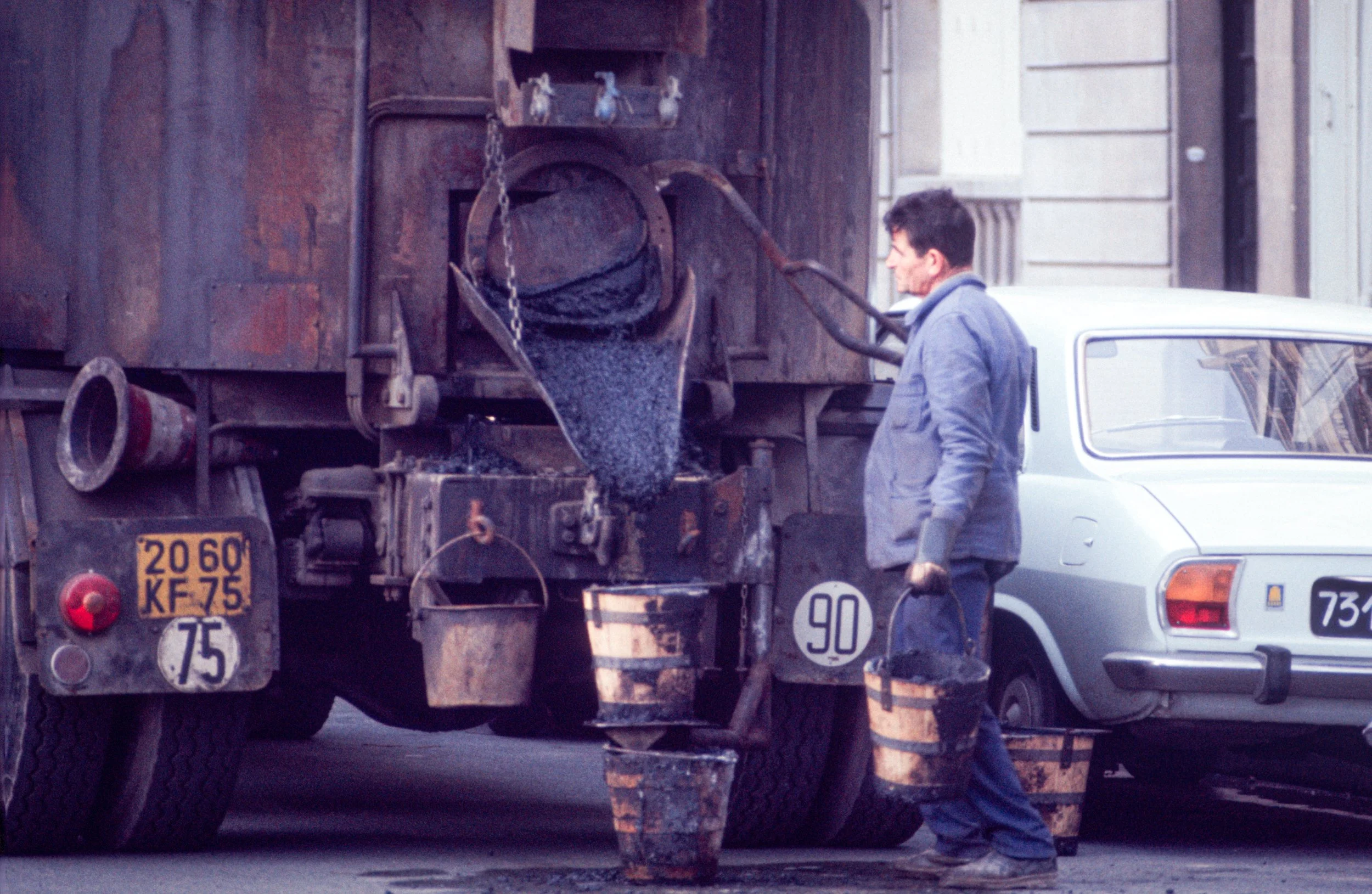 A man in a gray jacket collects asphalt from a mobile asphalt mixer truck into buckets on a city street.