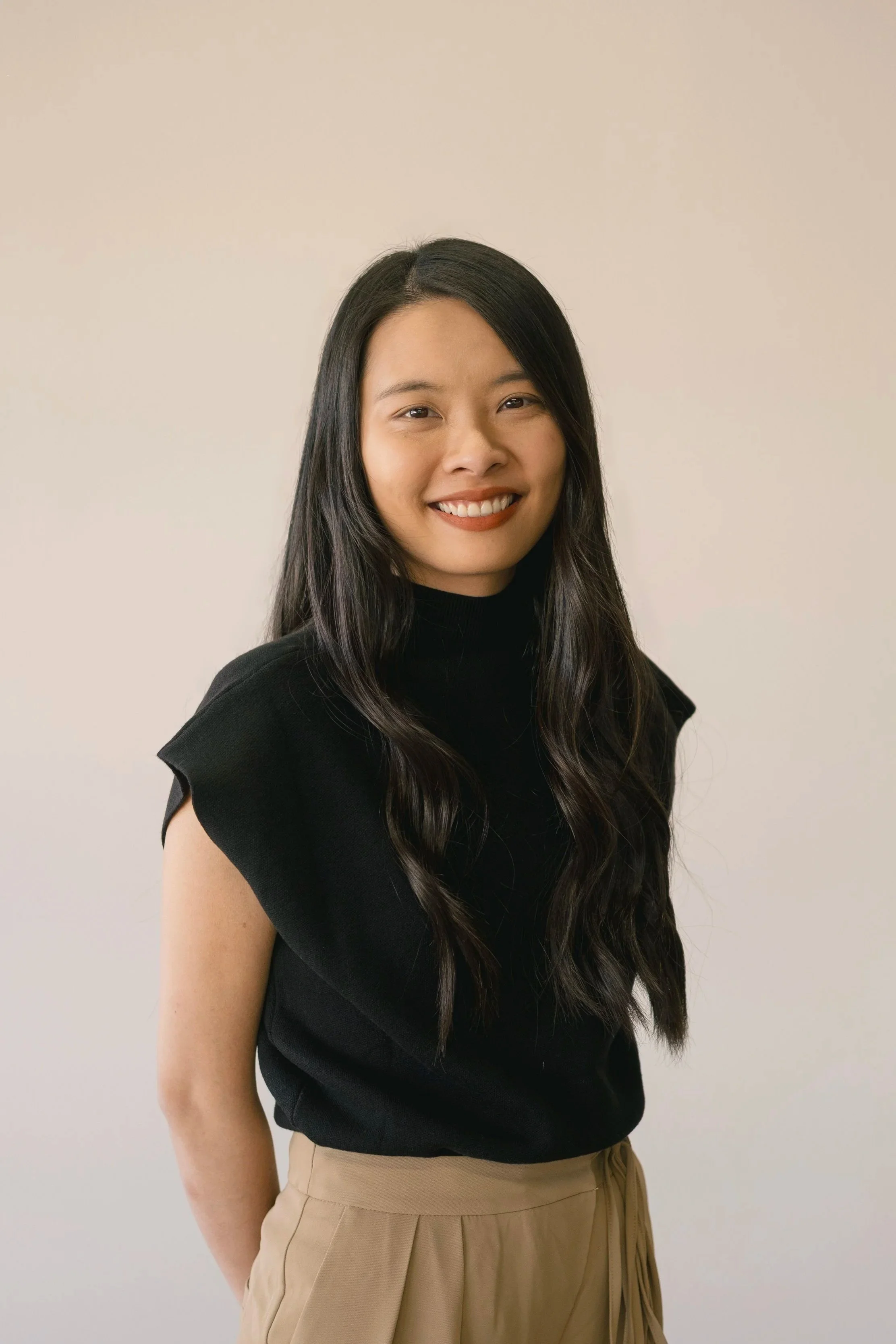 Portrait of a young woman with long dark hair, wearing a black top and beige pants, smiling against a plain light-colored background.