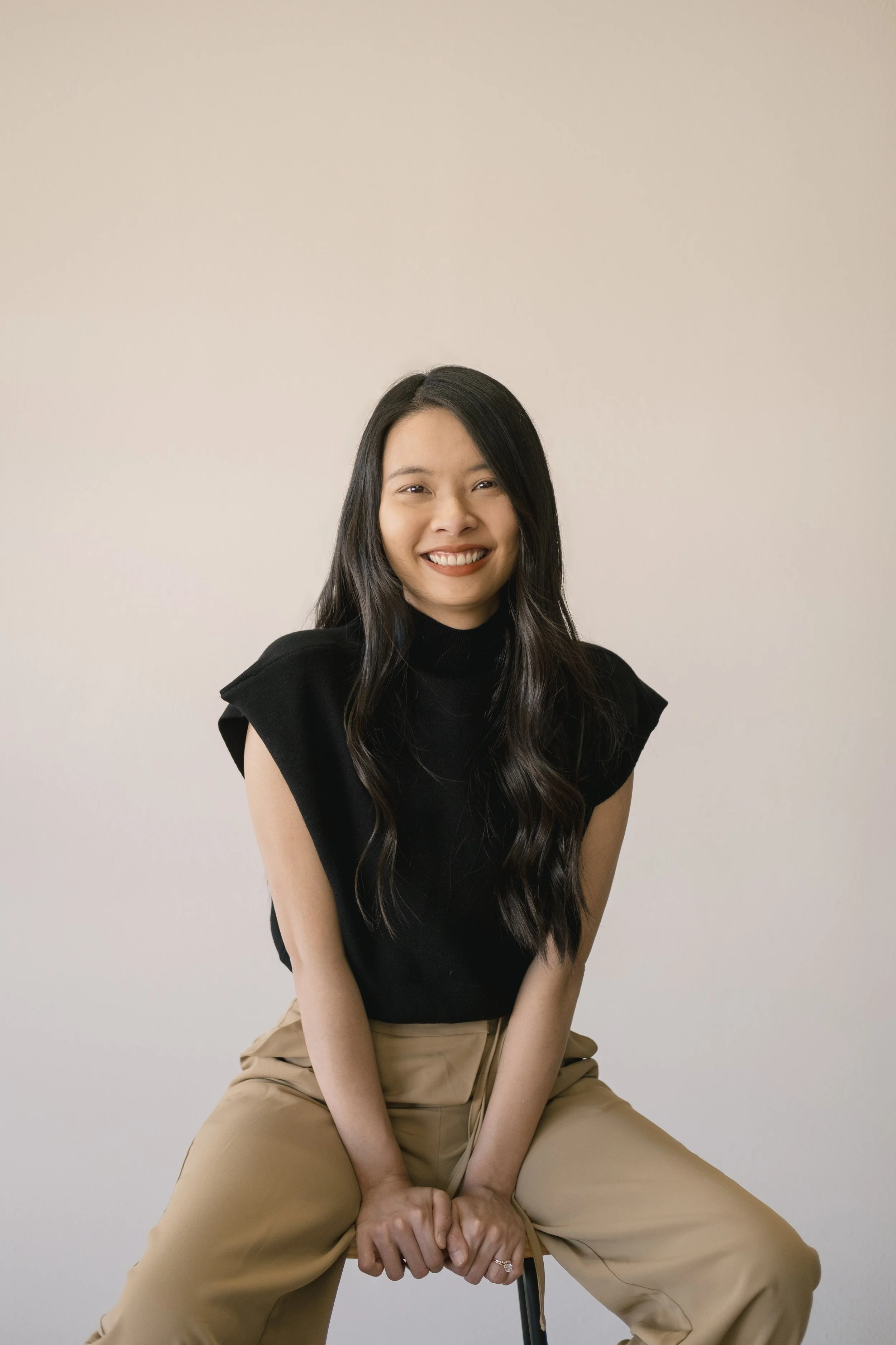 A young woman with long dark hair, wearing a black sleeveless top and beige pants, sitting on a chair against a plain light background, smiling and looking at the camera.