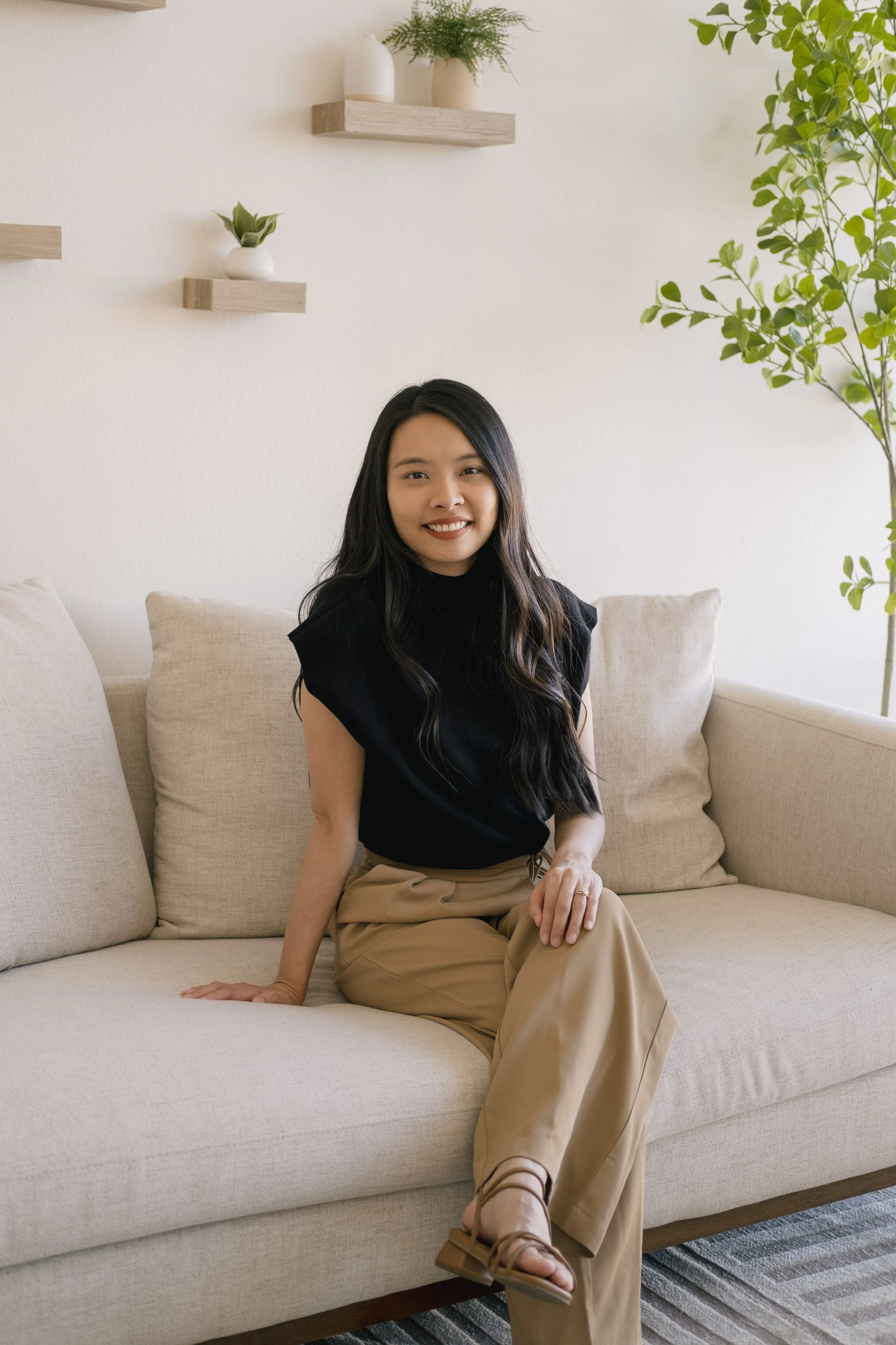 A woman with long black hair sitting on a light beige sofa in a room with a white wall. She is wearing a black top and tan pants, smiling at the camera. There are three small wooden shelves with white vases and plants on the wall behind her, and a large green plant on the right side of the image.
