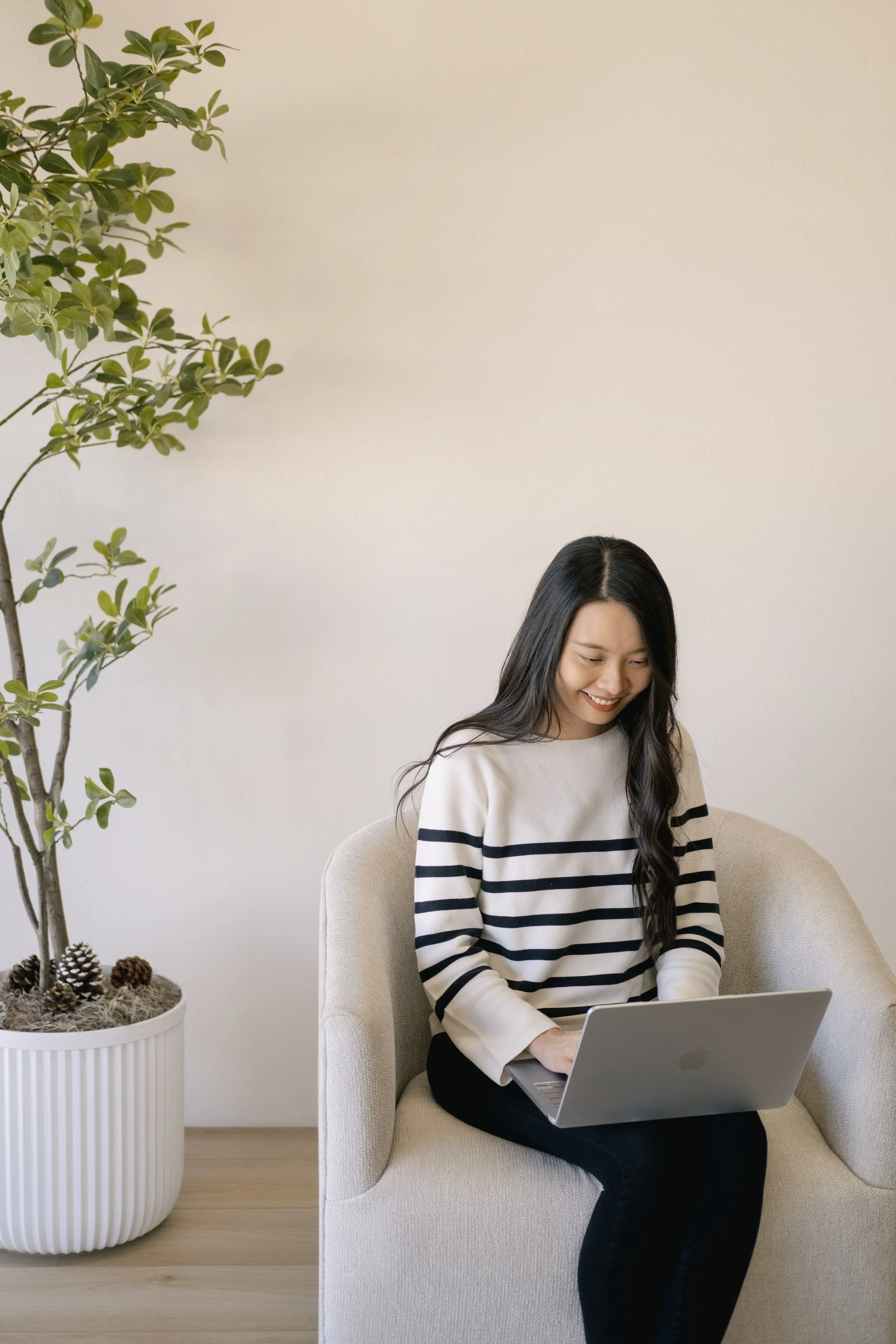 A woman sitting on a beige chair at a desk, using a silver laptop, with a potted plant with green leaves nearby, against a plain light-colored wall.