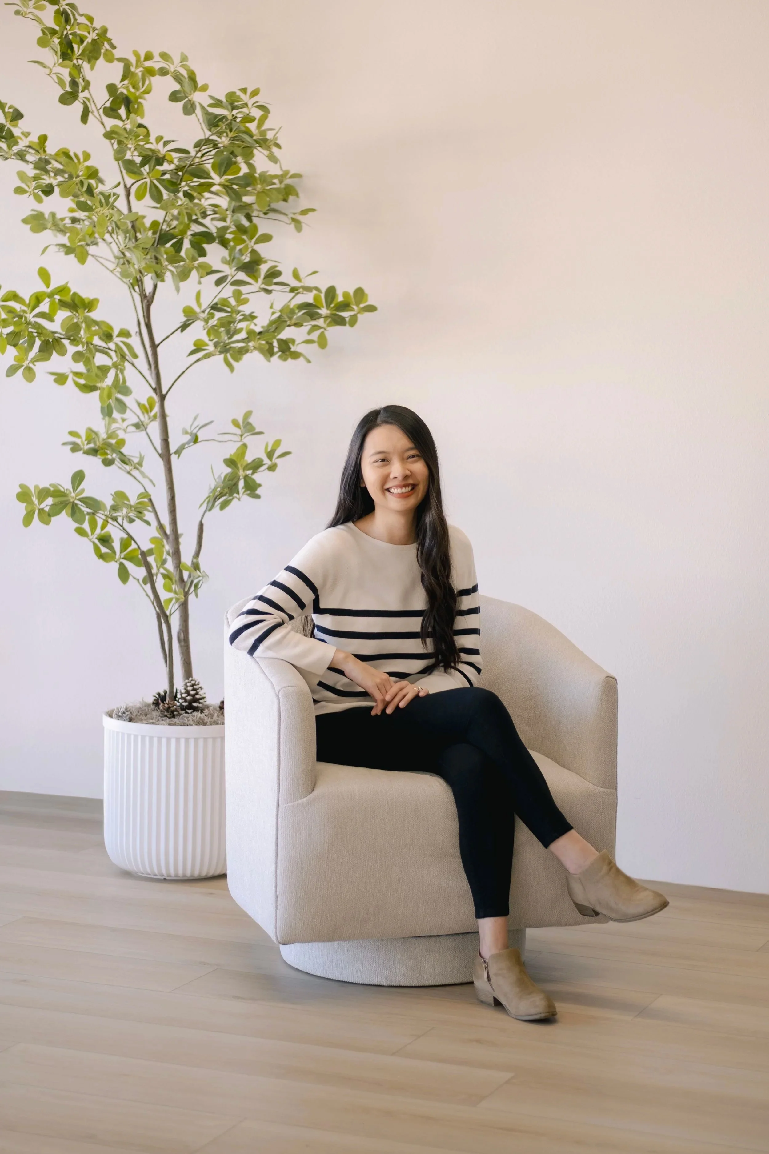 A woman sitting on a beige armchair next to a large potted plant with green leaves, smiling at the camera in a minimalistic room with light wood flooring and a white wall.