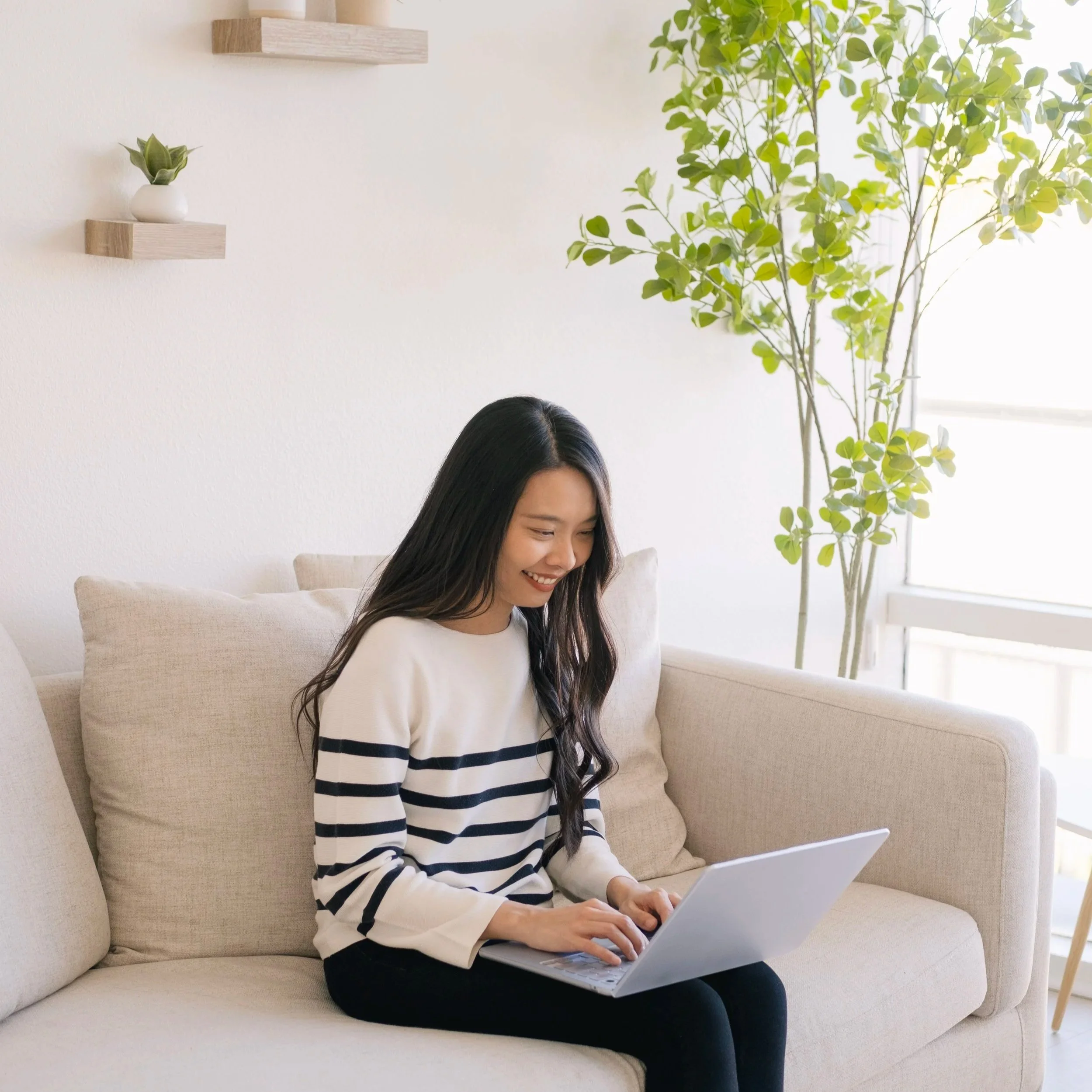A young woman with long dark hair sitting on a beige couch, smiling and using a silver laptop, in a bright, minimalistic living room with white walls, wooden shelves, a potted plant, and a large window.