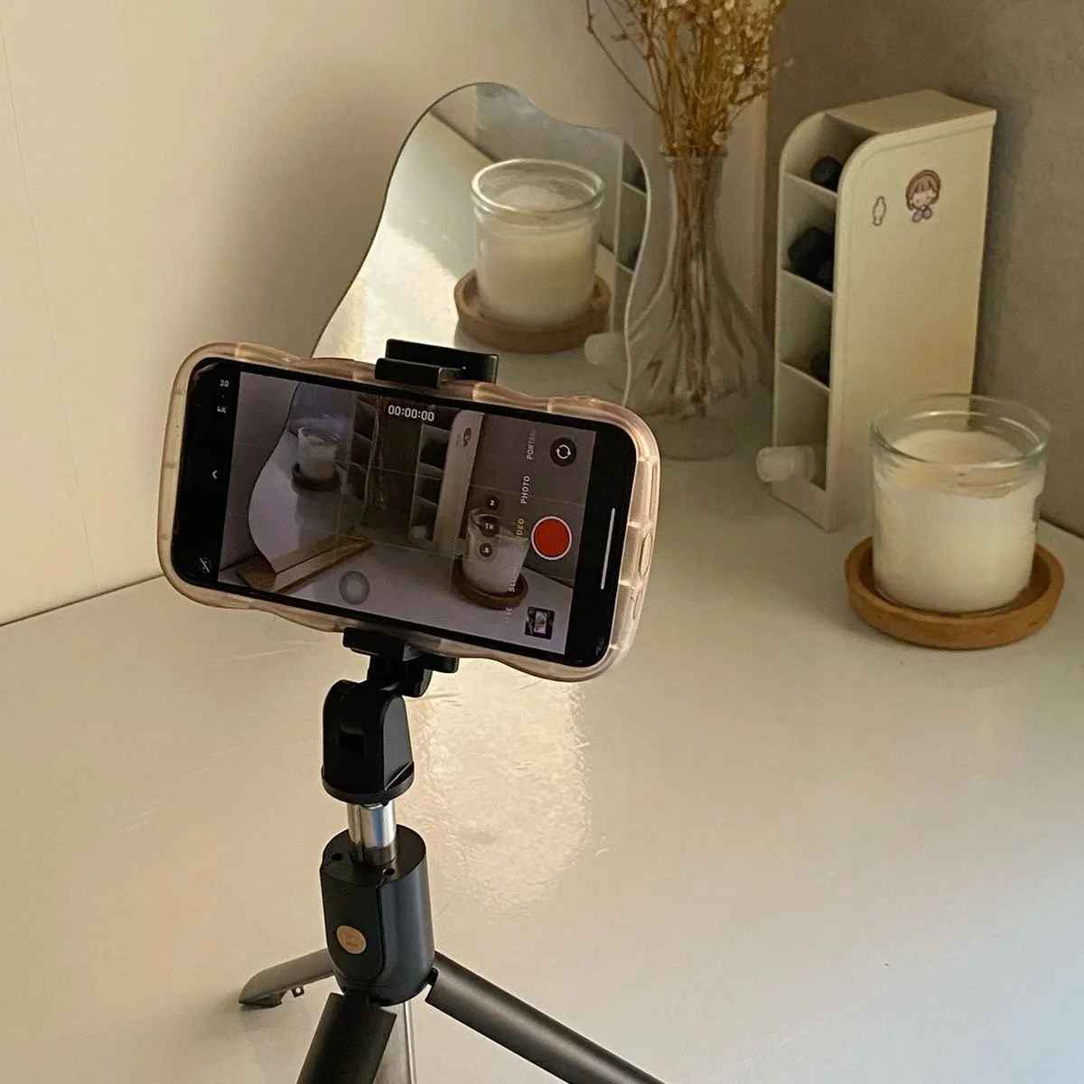 A smartphone on a small tripod capturing a scene with candles, dried flowers, and a small white shelf with compartments, all reflected in a mirror.