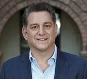 Portrait of a man in a blue jacket and white shirt, smiling, standing outdoors in front of a brick building.