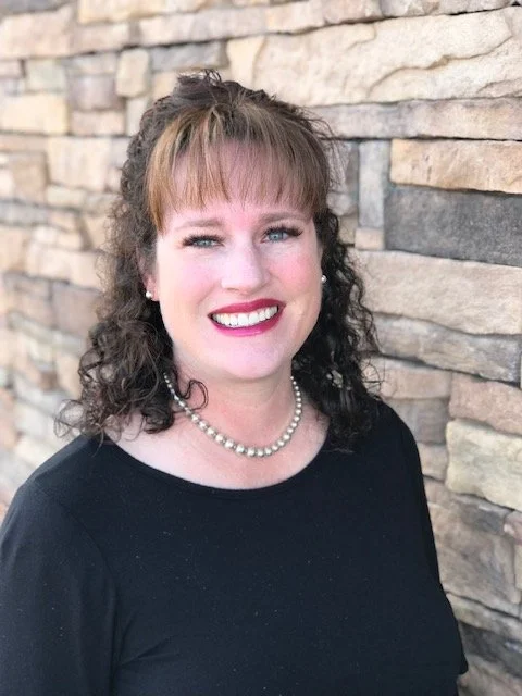 A woman with curly brown hair and bangs, wearing a pearl necklace and a black top, smiling in front of a stone wall.