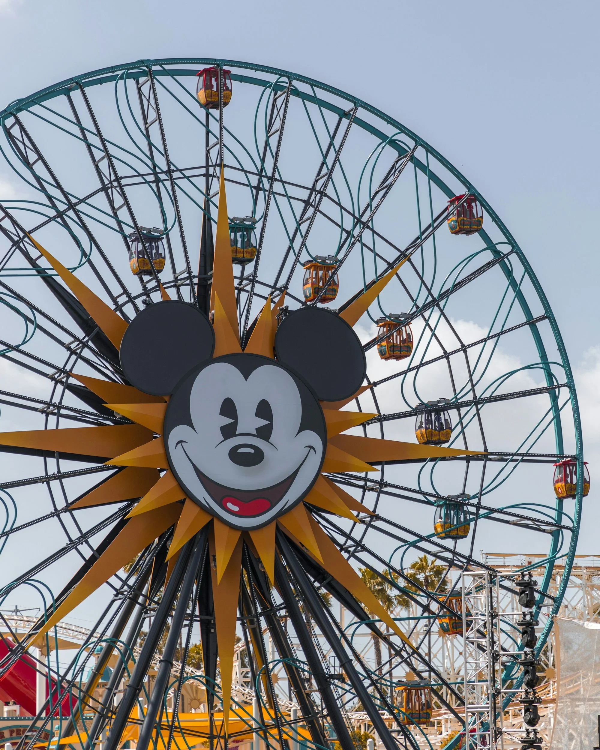 A large Ferris wheel with a Mickey Mouse face and sun rays at the center, featuring several gondolas and a clear sky in the background.