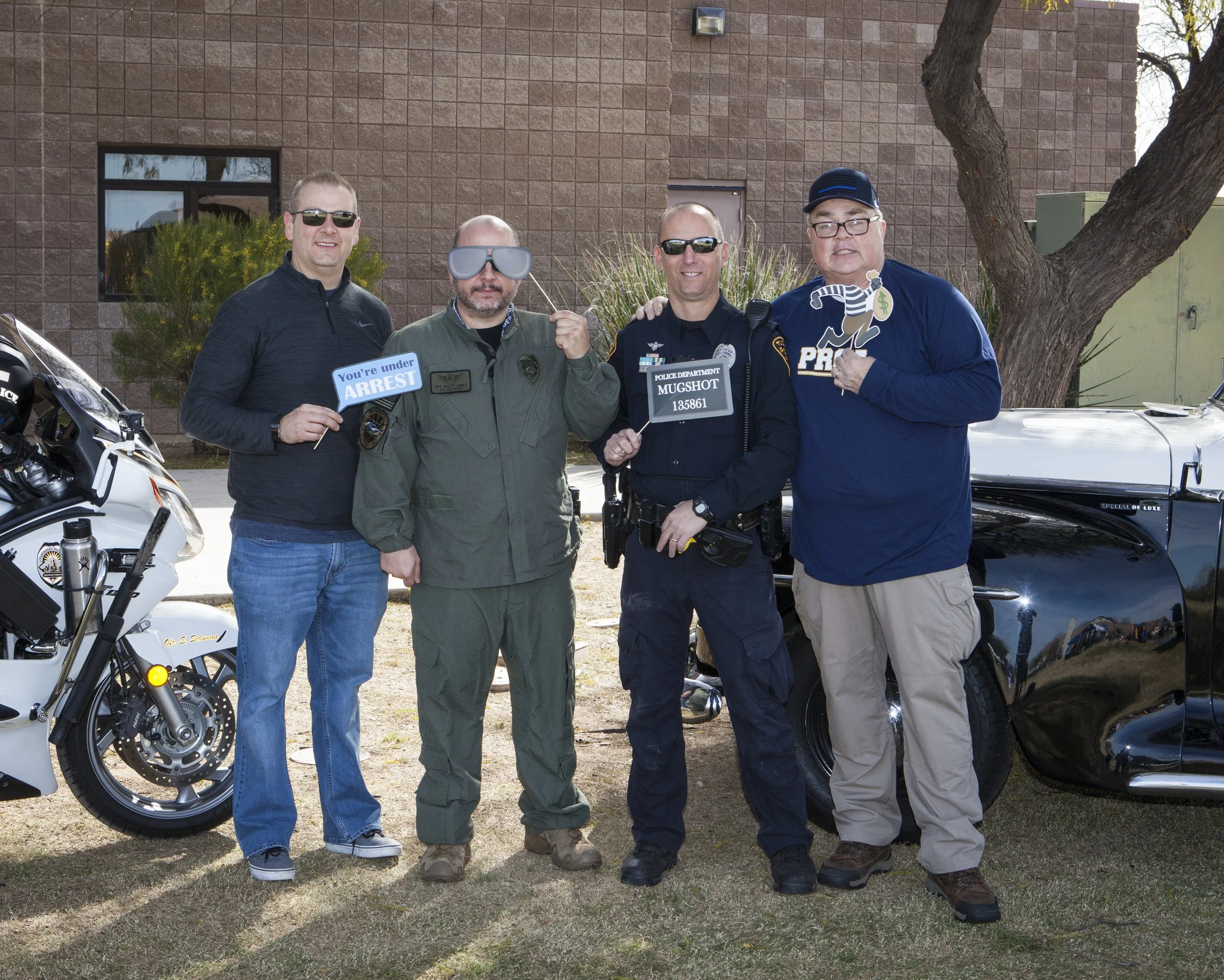 Four men standing outdoors in front of a building, holding signs related to police, with police motorcycles and cars nearby. One man wears a police uniform, others are casually dressed.