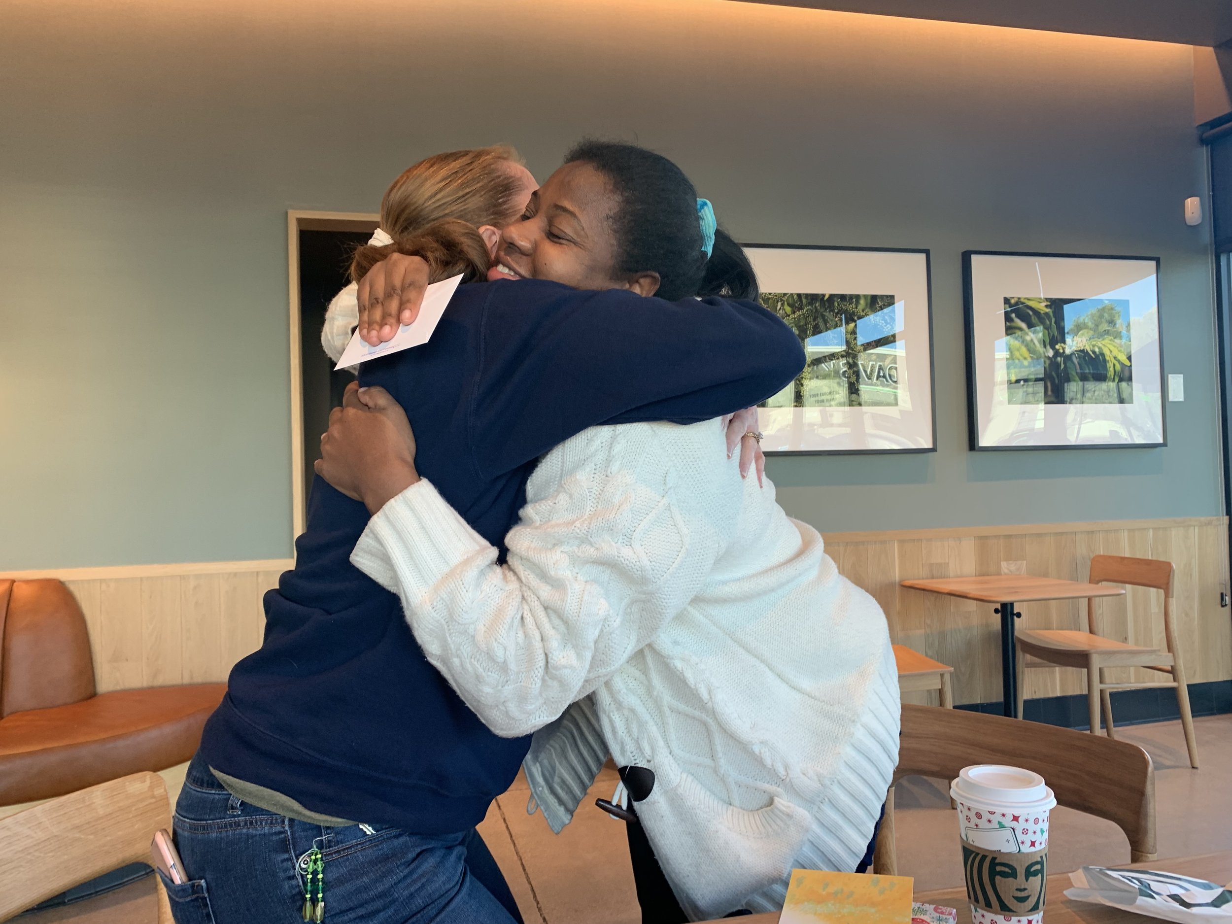 Two women hugging tightly inside a cafe, with smiling faces. One woman has blonde hair, wearing a light-colored sweater, while the other has dark hair with a bright blue hair tie, wearing a navy blue top.