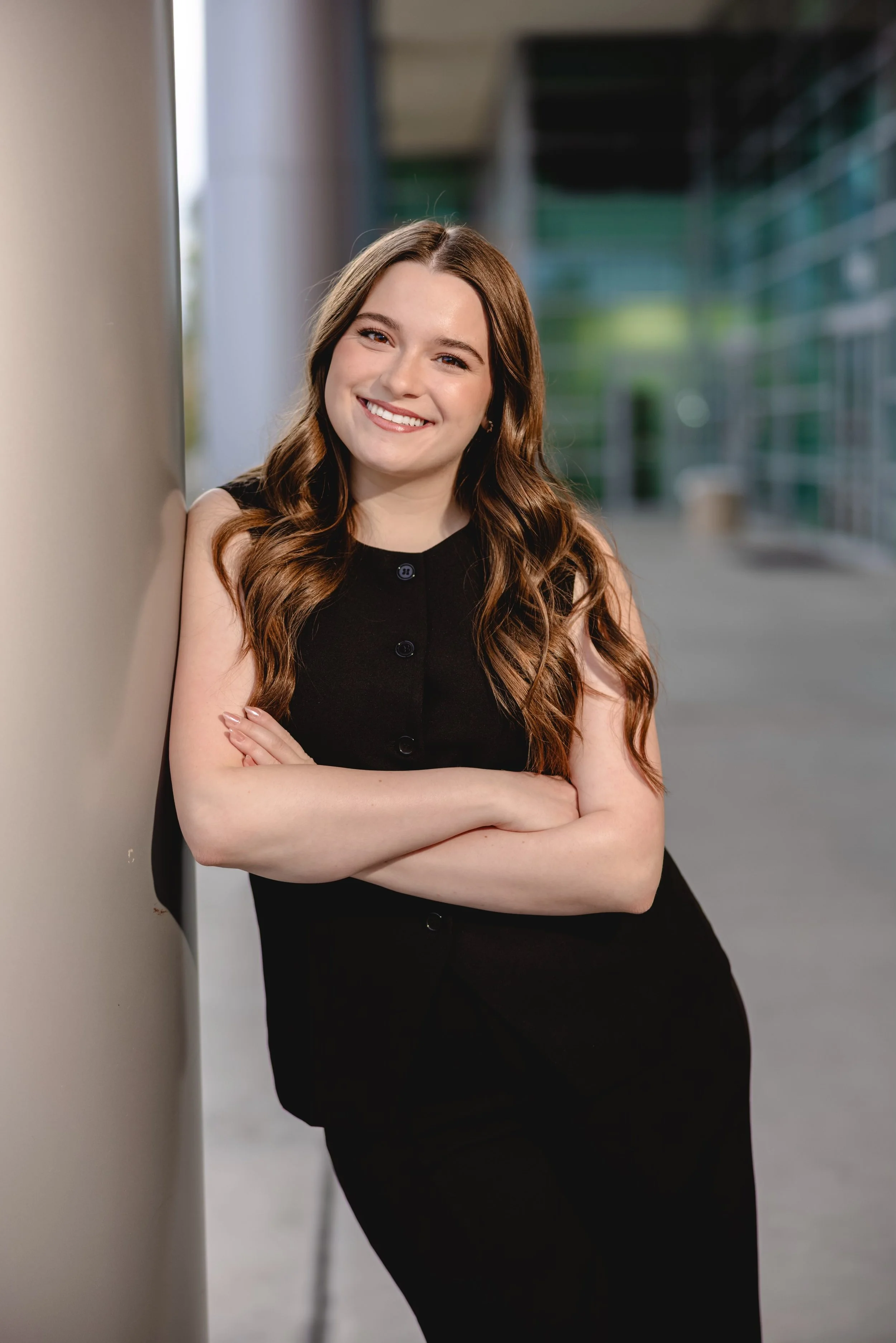 A young woman with long wavy brown hair, smiling, leaning against a wall with arms crossed, outdoors in an urban setting.