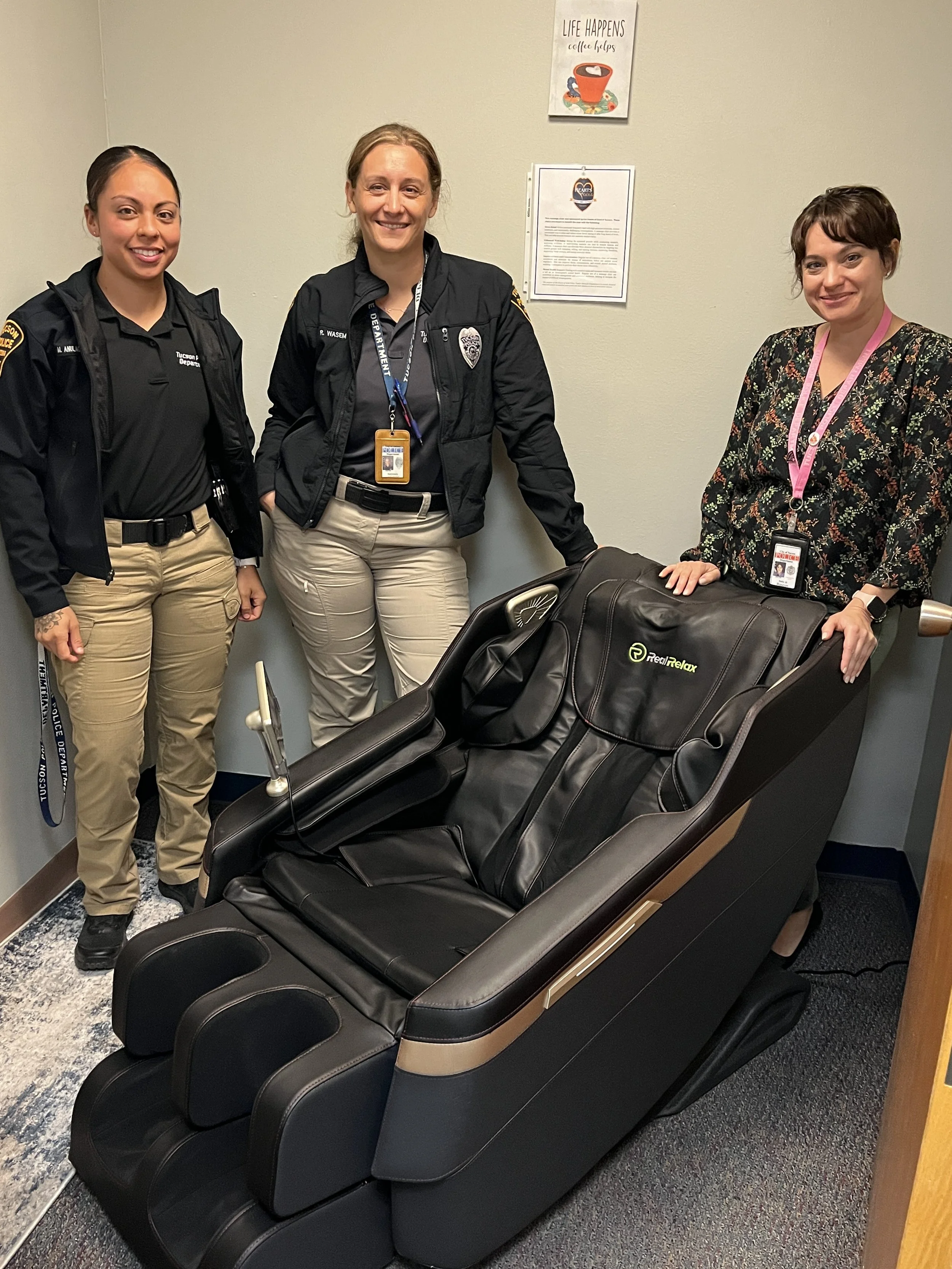 Three women standing around a massage chair in an office or medical setting. The women are smiling, and the massage chair is black with tan accents.