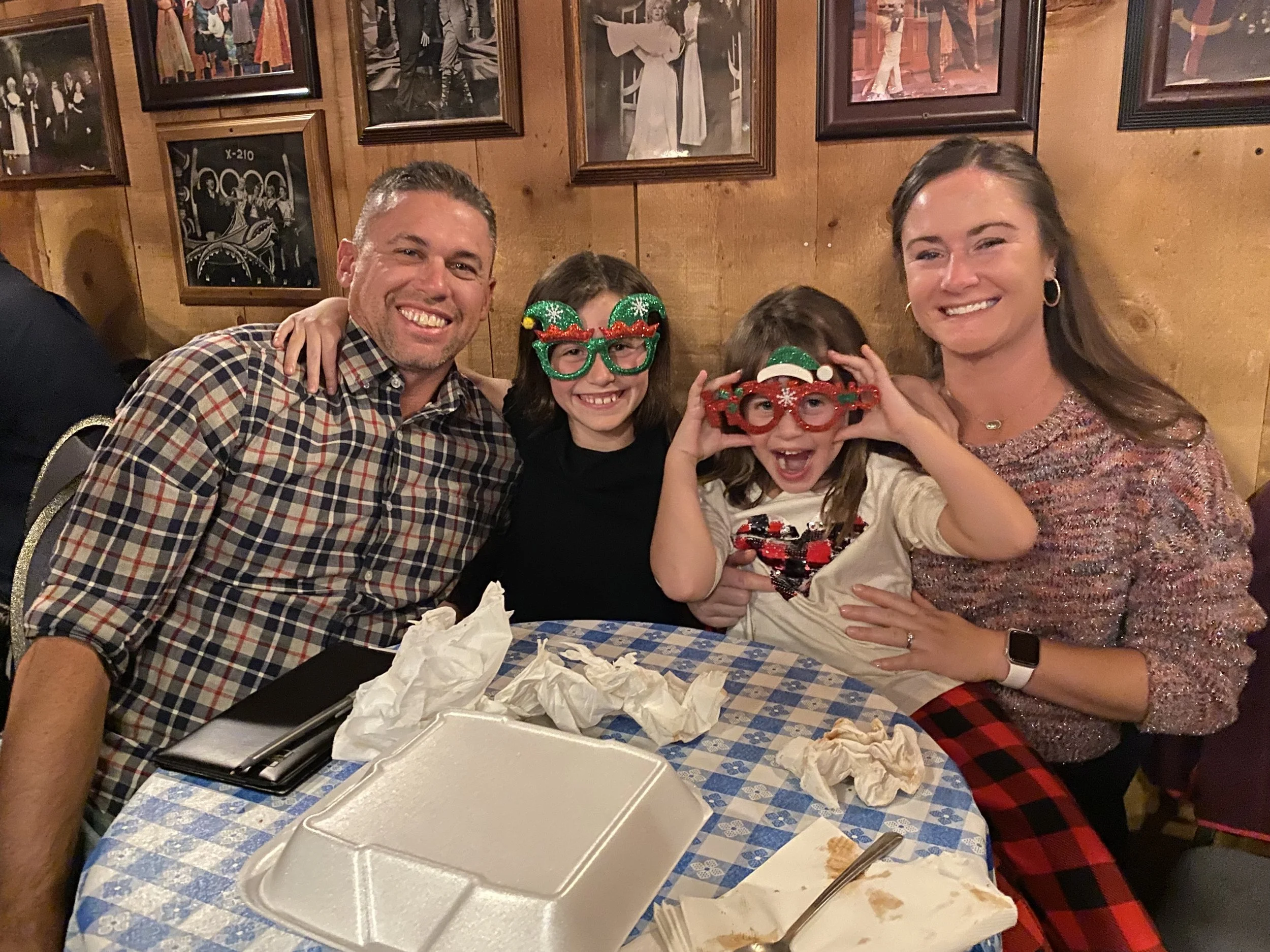A group of four people, two adults and two children, sitting at a table in a restaurant, smiling at the camera. The children are wearing festive Christmas glasses, and the table has crumpled napkins and leftover food.