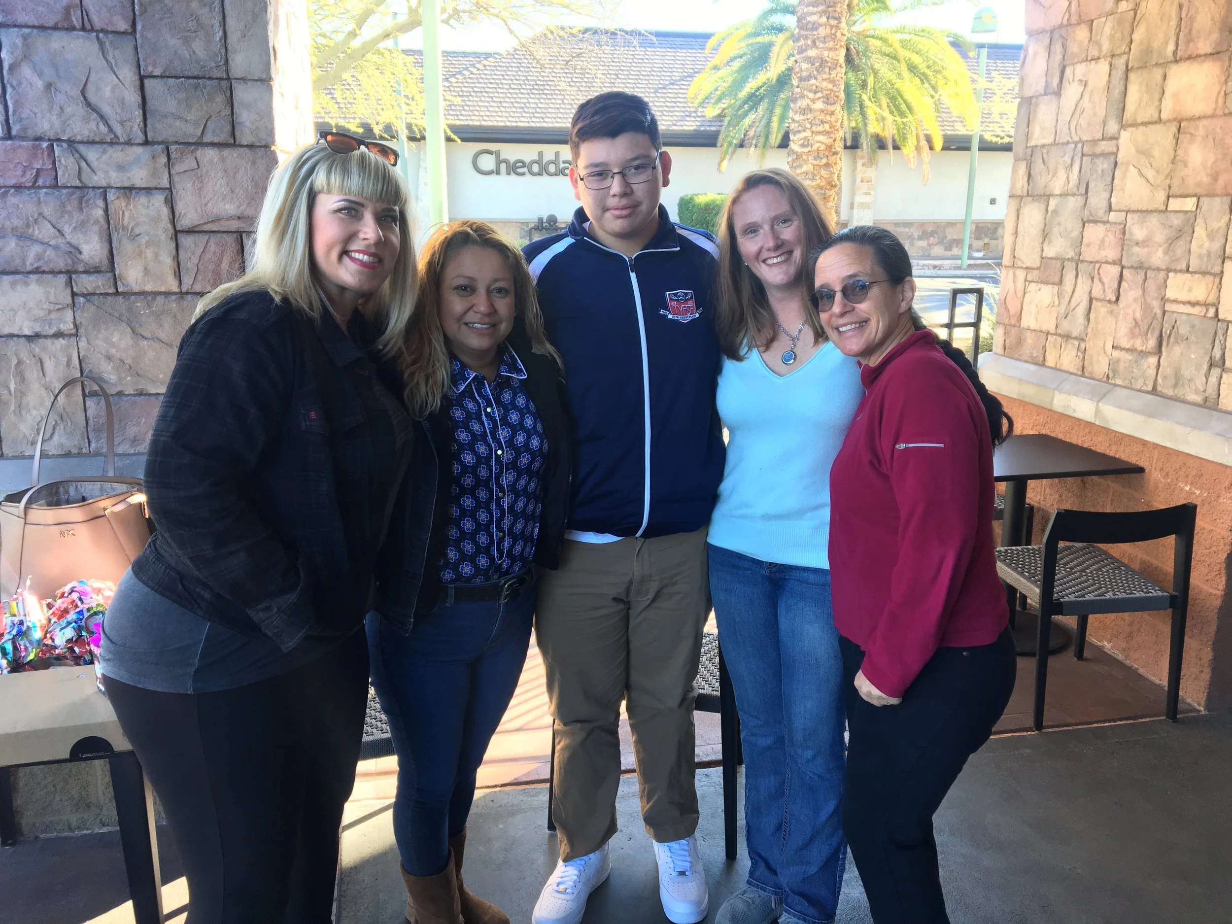 Group of five women and one young man standing outdoors, smiling for a photo, with a palm tree and restaurant sign in the background.