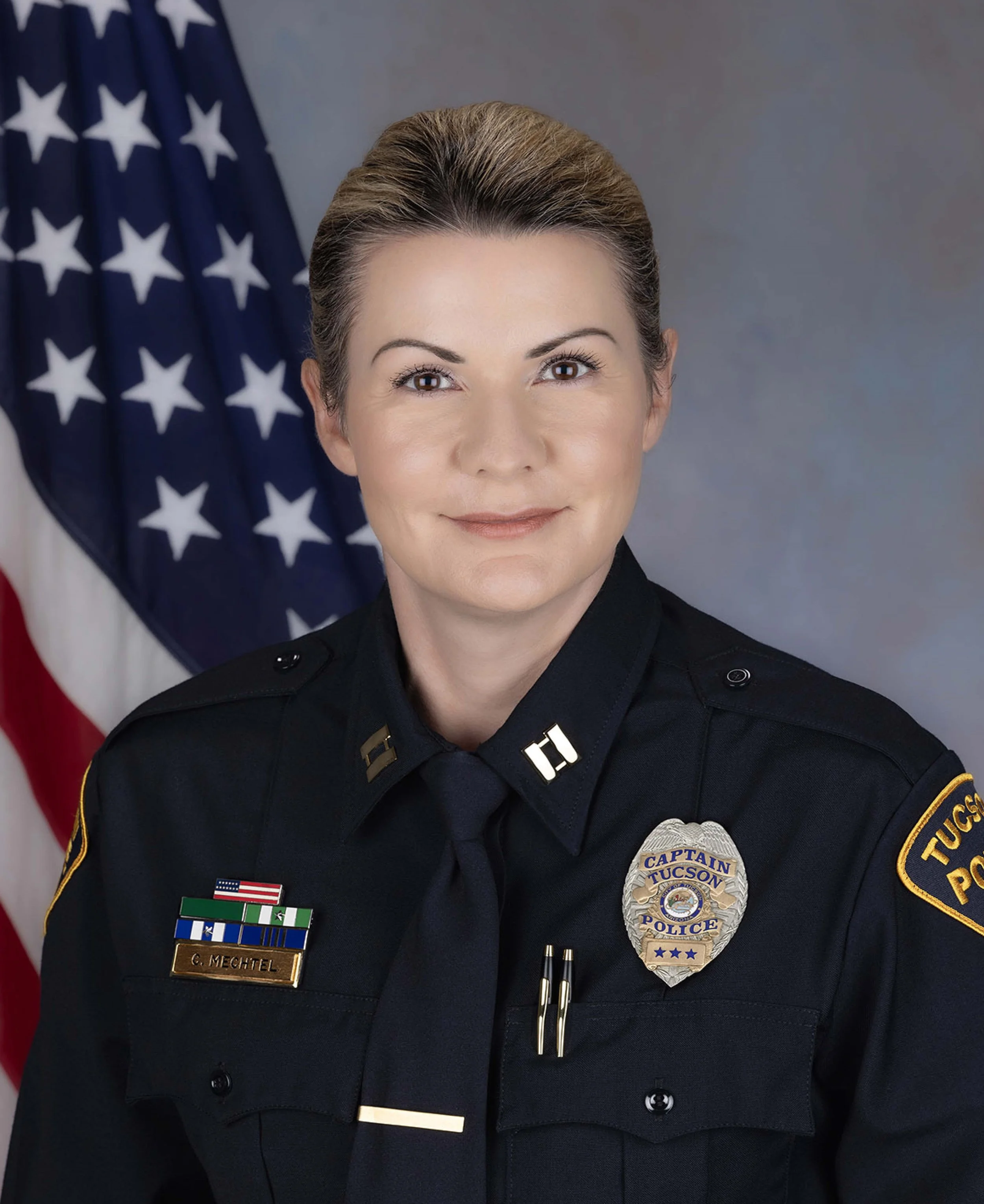 A female police officer in uniform with a badge, standing in front of an American flag.