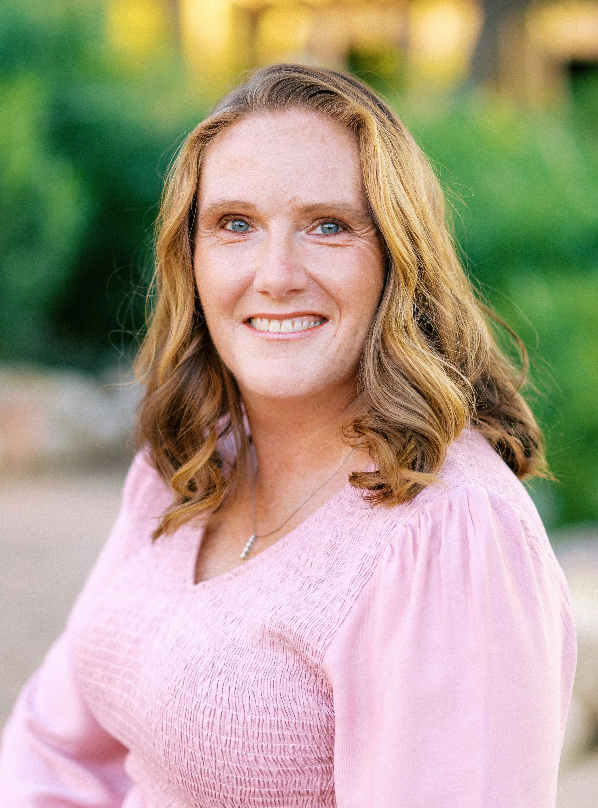 A woman with curly, shoulder-length light brown hair, blue eyes, and freckles, wearing a pink blouse and a necklace, smiling outdoors with a blurred background of greenery.