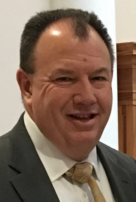 A middle-aged man with short dark hair, smiling, wearing a suit and tie, inside a room with wooden furniture visible in the background.