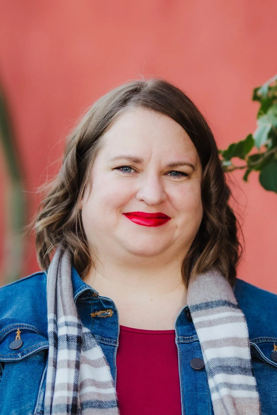 A woman with shoulder-length wavy brown hair, wearing bright red lipstick, a denim jacket, a magenta shirt, and a gray plaid scarf, posing outdoors with a pink background and some green plants visible on the right side.