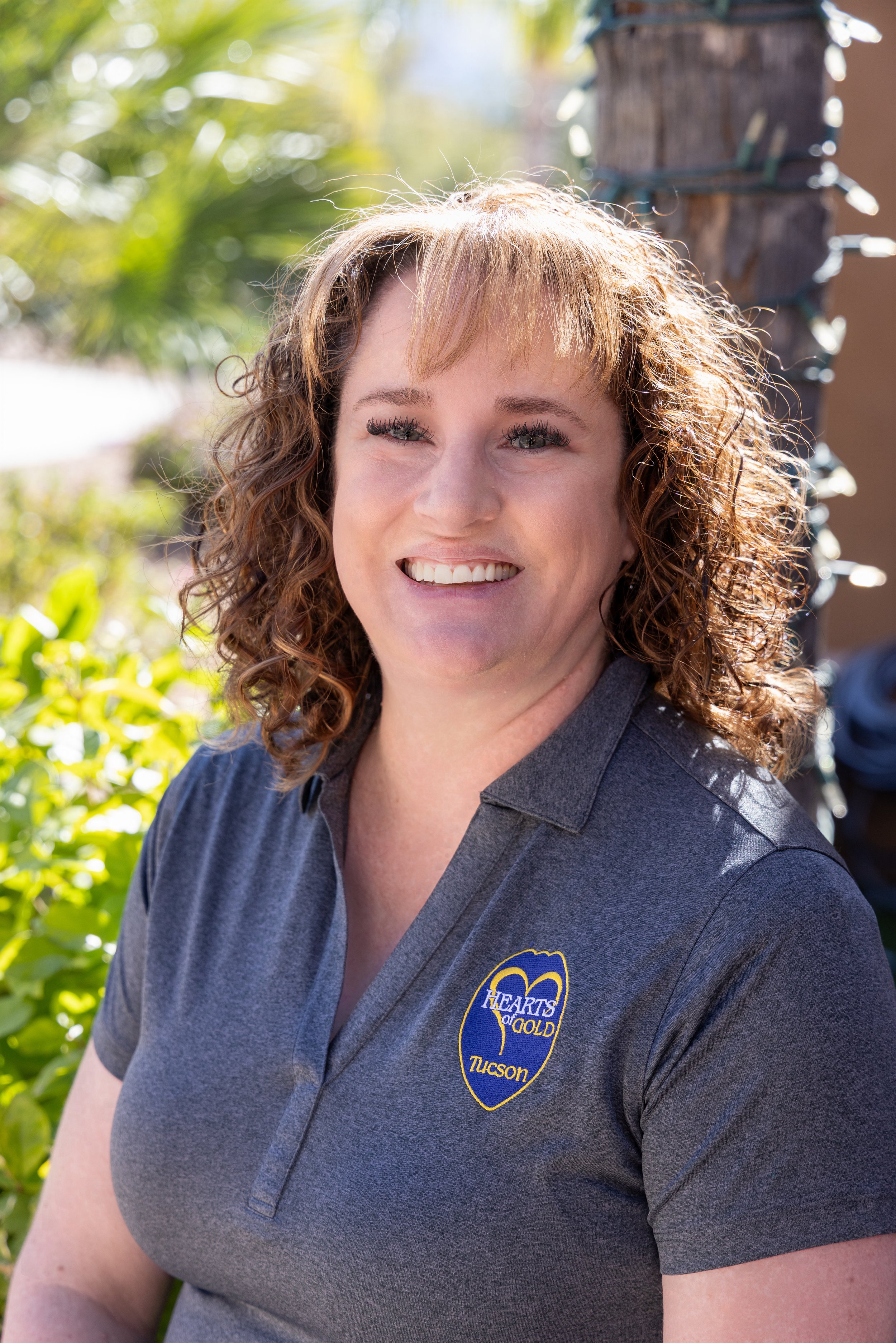A woman with curly brown hair and bangs, wearing a pearl necklace and a black top, smiling in front of a stone wall.