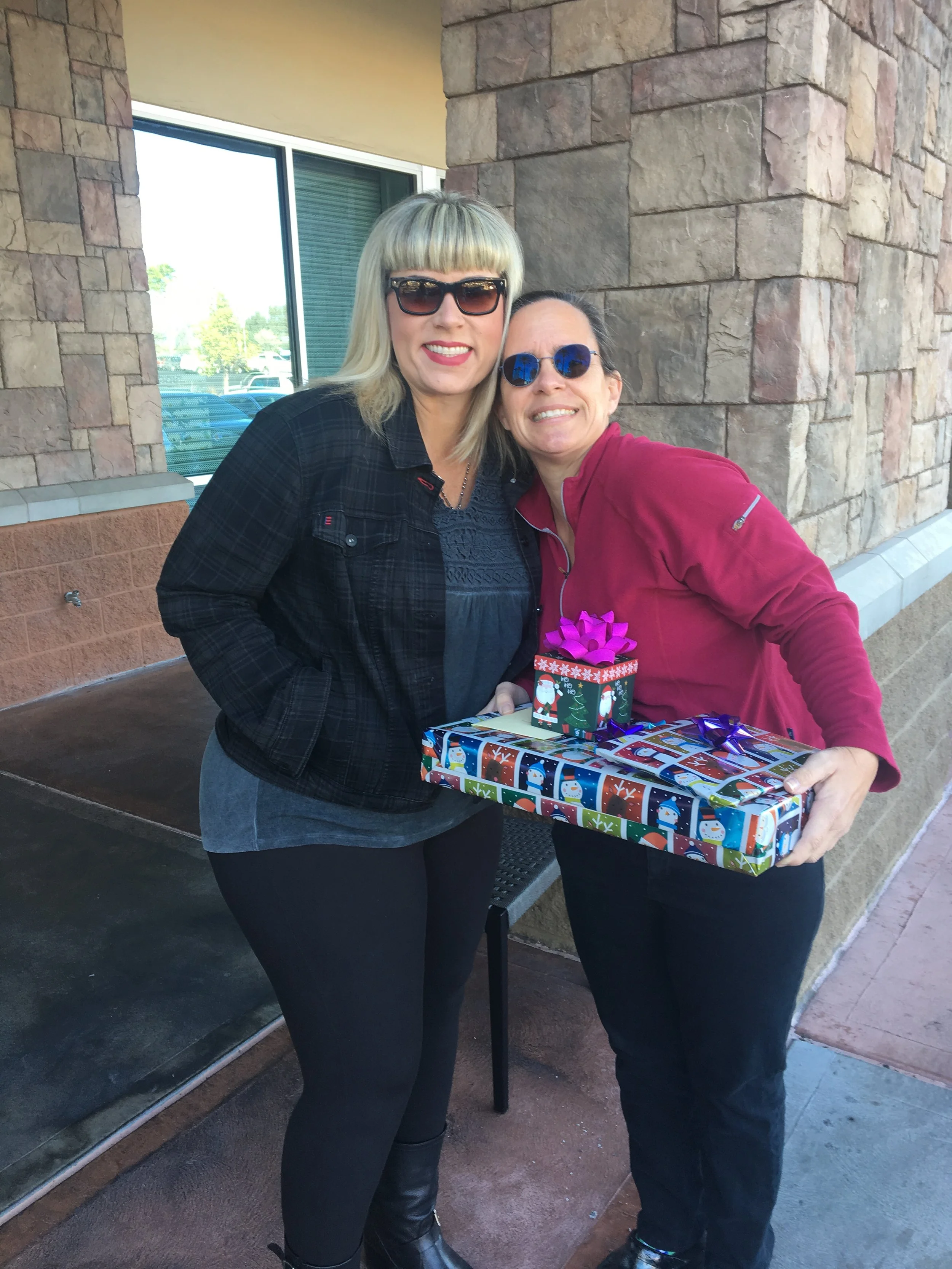 Two women standing outside near a brick wall, holding a wrapped gift with Christmas-themed paper and a smaller gift on top, both smiling at the camera.