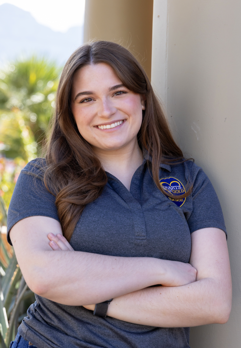 A young woman with long wavy brown hair, smiling, leaning against a wall with arms crossed, outdoors in an urban setting.