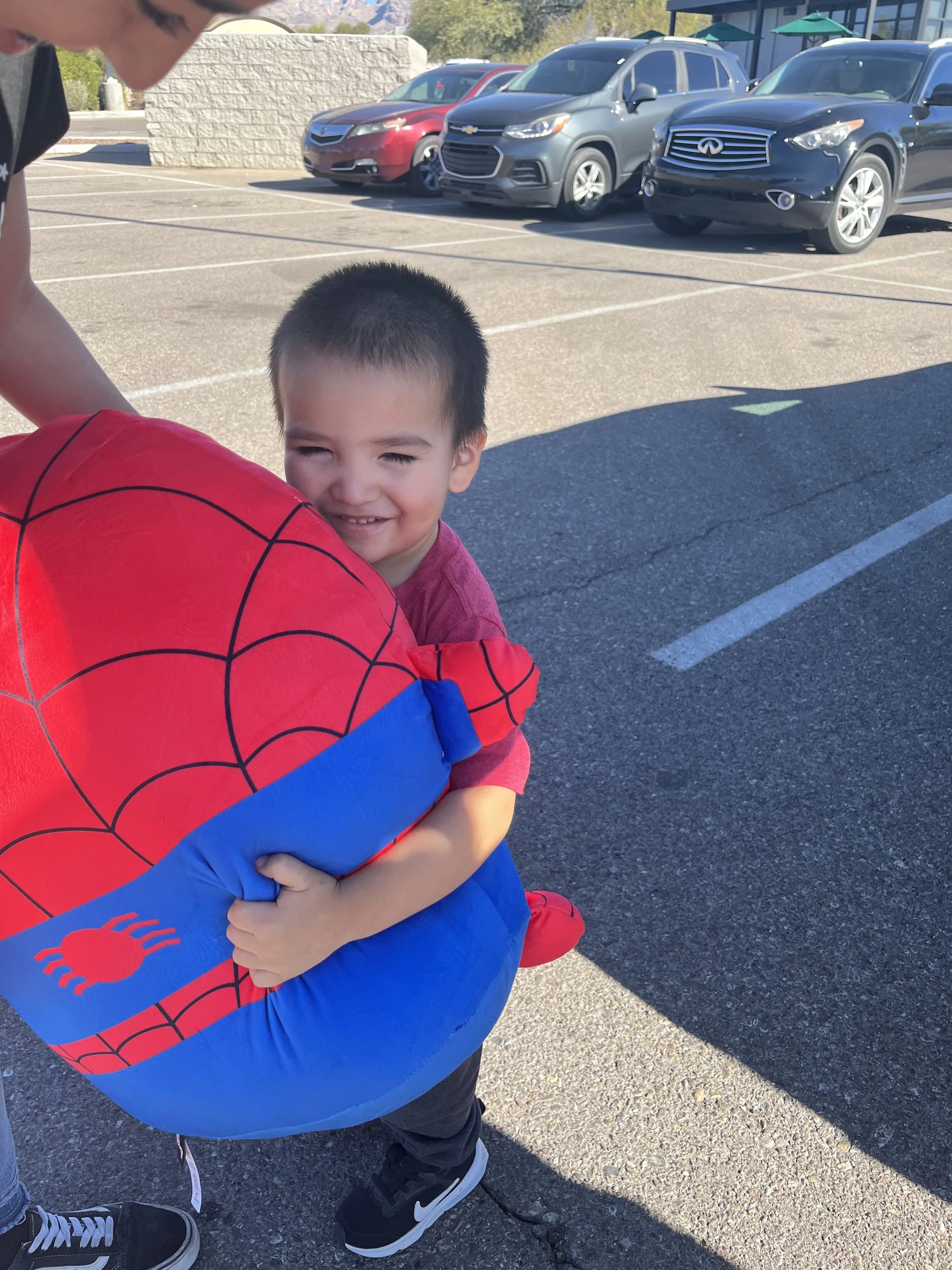 A young boy in a red shirt embraces a large Spider-Man themed inflatable of Spider-Man's head, smiling in a parking lot with cars in the background.