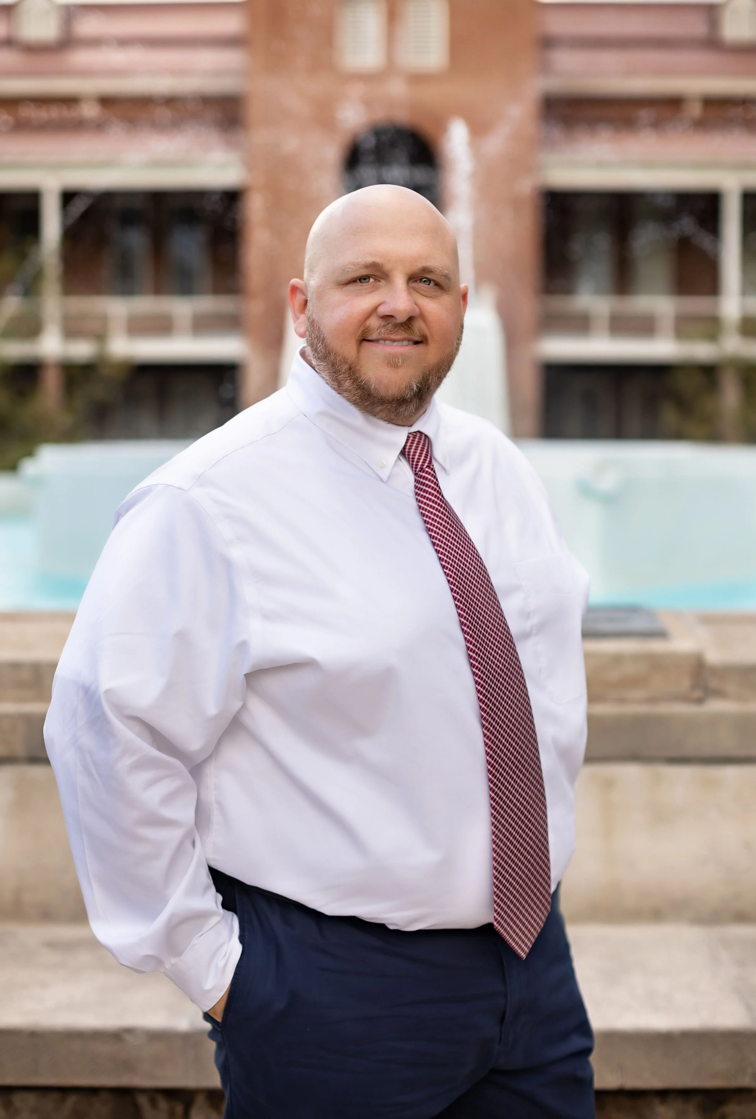 A man in a white dress shirt and red patterned tie standing outdoors in front of a fountain with a historic brick building in the background.
