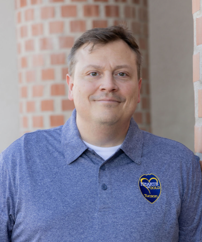 A man in a black suit, white shirt, and blue polka-dot tie seated against a wooden wall.