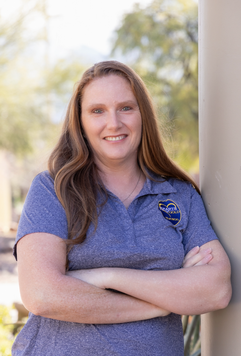 A woman with curly, shoulder-length light brown hair, blue eyes, and freckles, wearing a pink blouse and a necklace, smiling outdoors with a blurred background of greenery.