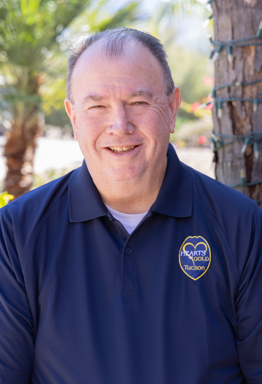 A middle-aged man with short dark hair, smiling, wearing a suit and tie, inside a room with wooden furniture visible in the background.