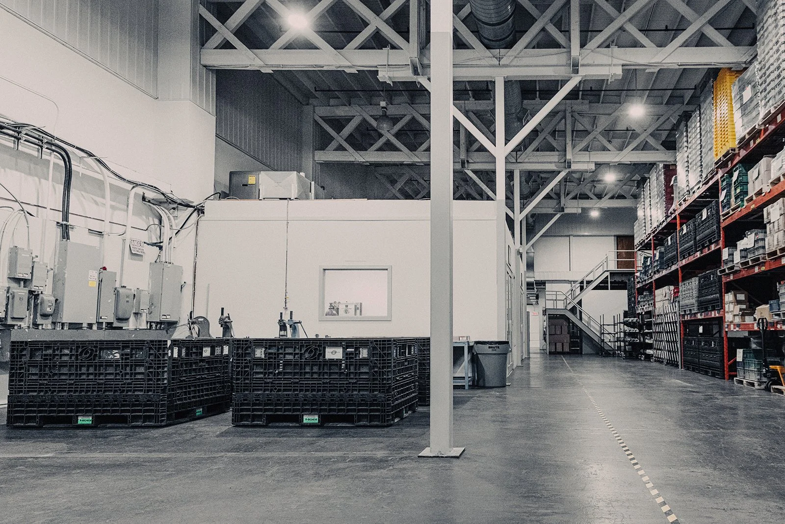 Empty warehouse aisle with industrial shelves, black plastic crates, and a staircase in the background.