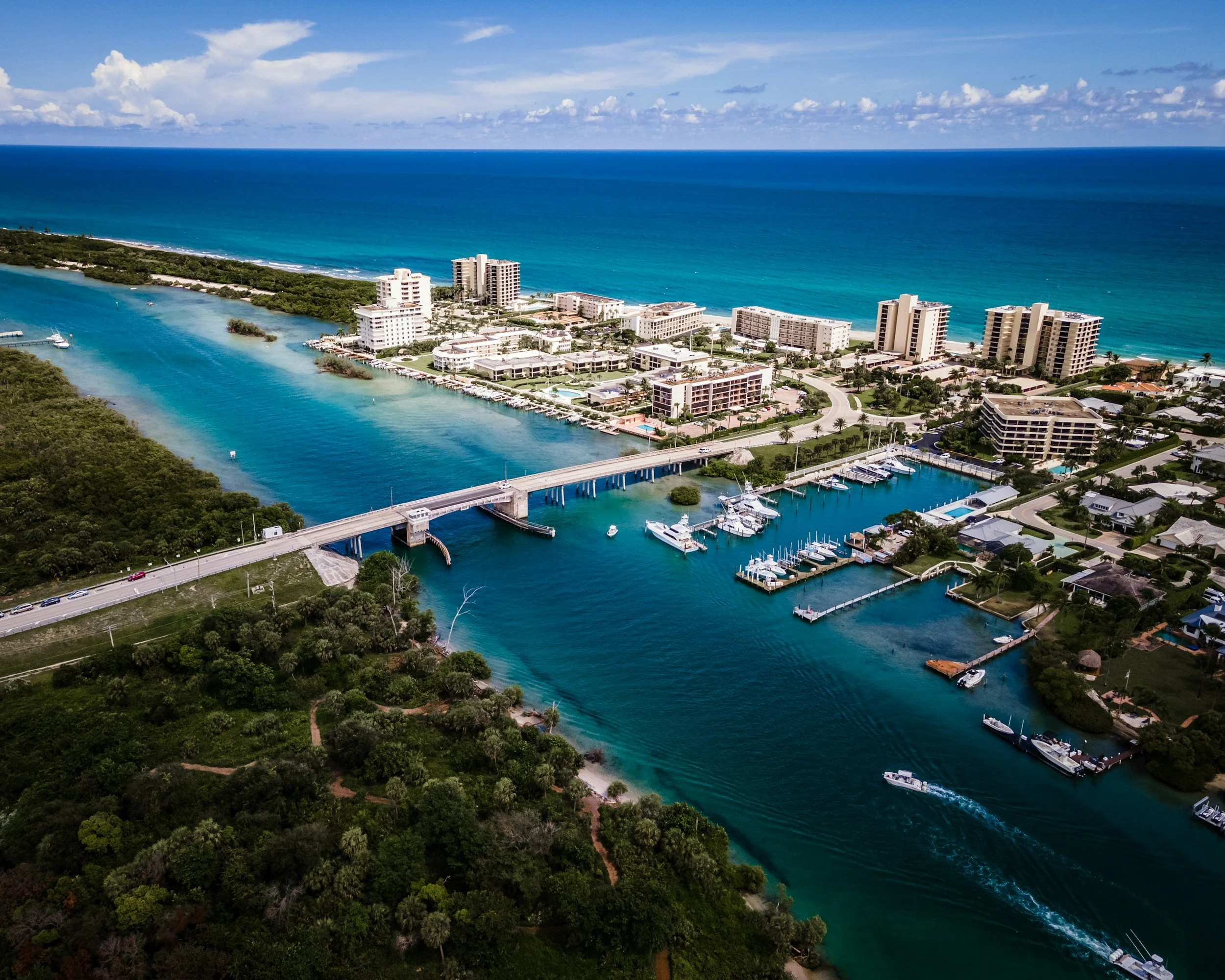 Aerial view of a coastal city with high-rise buildings, a marina with boats, a bridge over a waterway, lush greenery, and the ocean in the background under a partly cloudy sky.