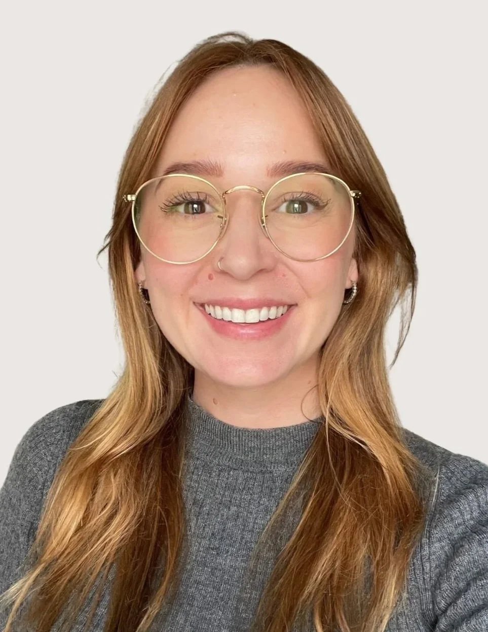 A woman with long reddish-brown hair, wearing large round eyeglasses and a gray sweater, smiling against a plain white background.