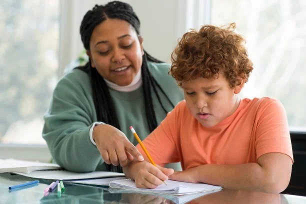 A woman helping a young boy with his homework at a desk, with papers and colored pencils on the table.