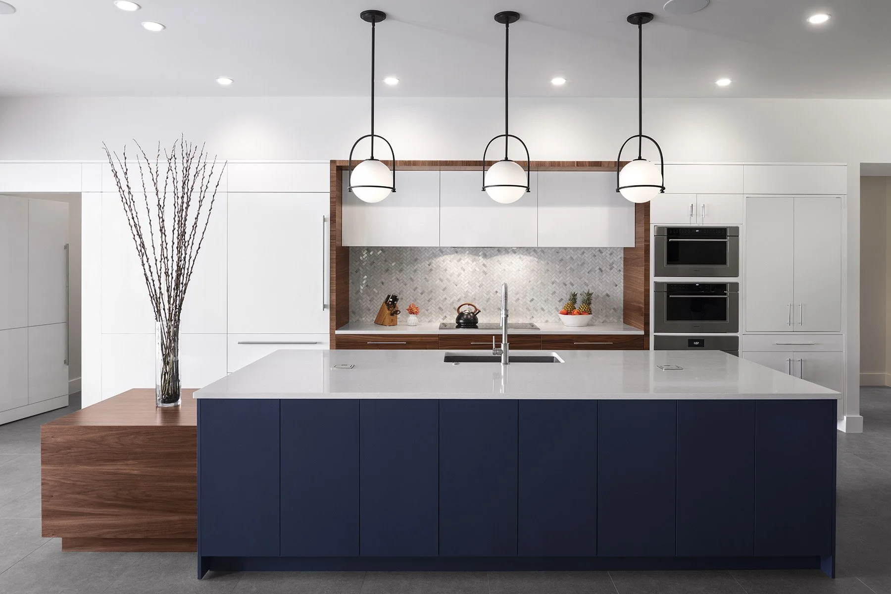 Modern kitchen with white upper cabinets, a central island with a white countertop and blue base, three hanging pendant lights, a stainless steel sink, a marble backsplash, and built-in double ovens. A tall vase with branches is on a wooden side table.