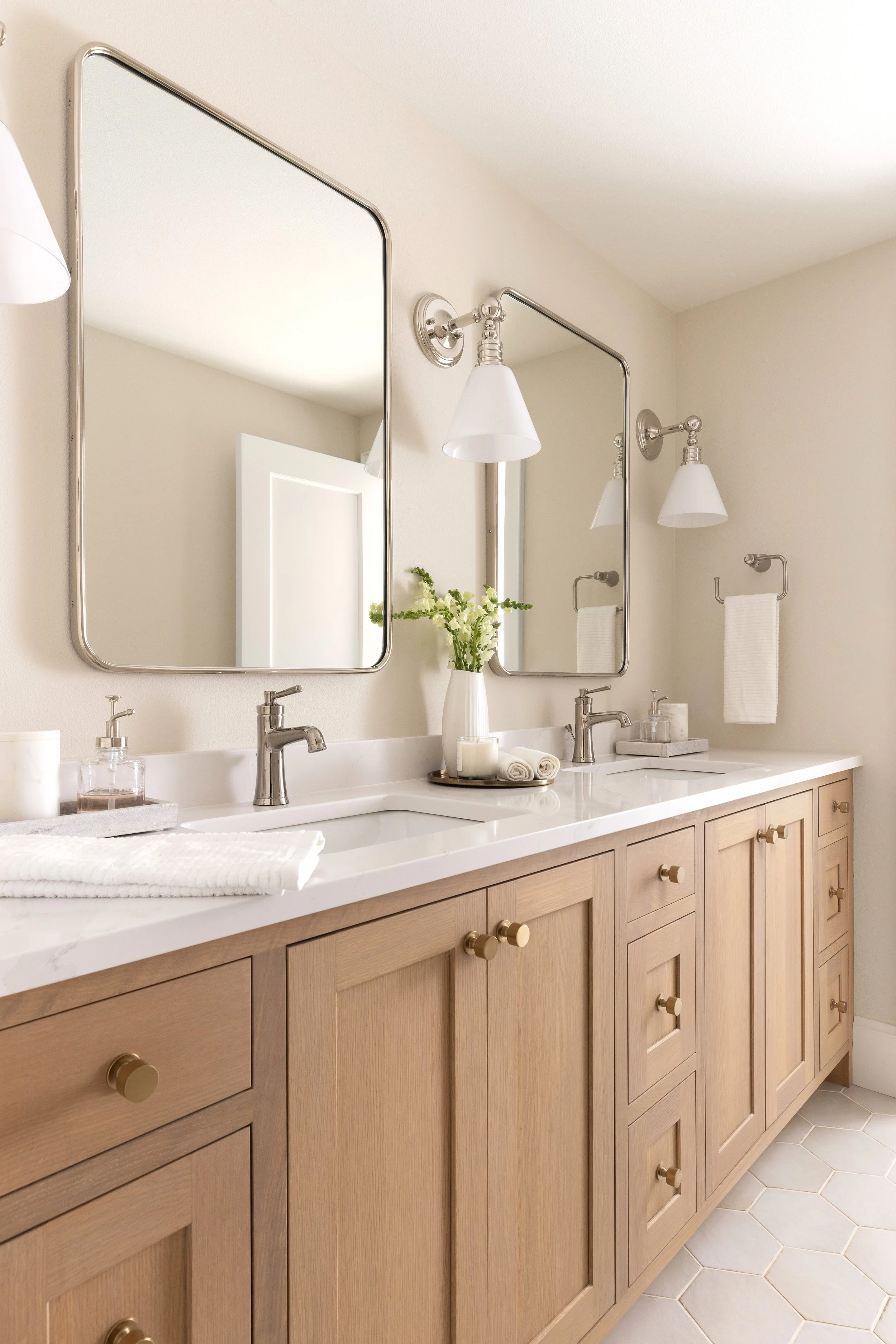 A modern bathroom vanity with a white countertop, dual sinks, and wooden cabinets with brass knobs. Two large rectangular mirrors are mounted above the sinks. Decorative elements include a vase with flowers, candles, and rolled towels. The bathroom features wall-mounted sconces with white shades and a hexagonal tiled floor.
