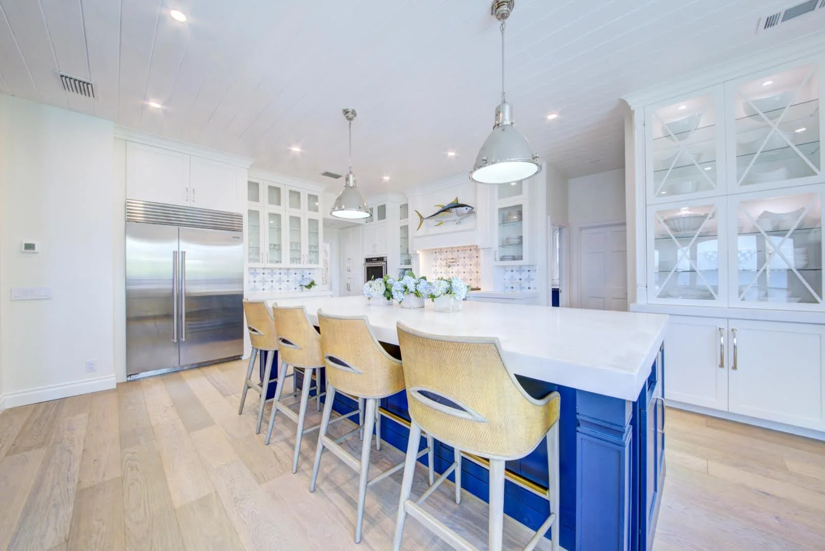 Bright white kitchen with a blue island, six yellow chairs, white cabinetry, a large stainless steel refrigerator, and nautical decor including fish wall art and a coral backsplash.