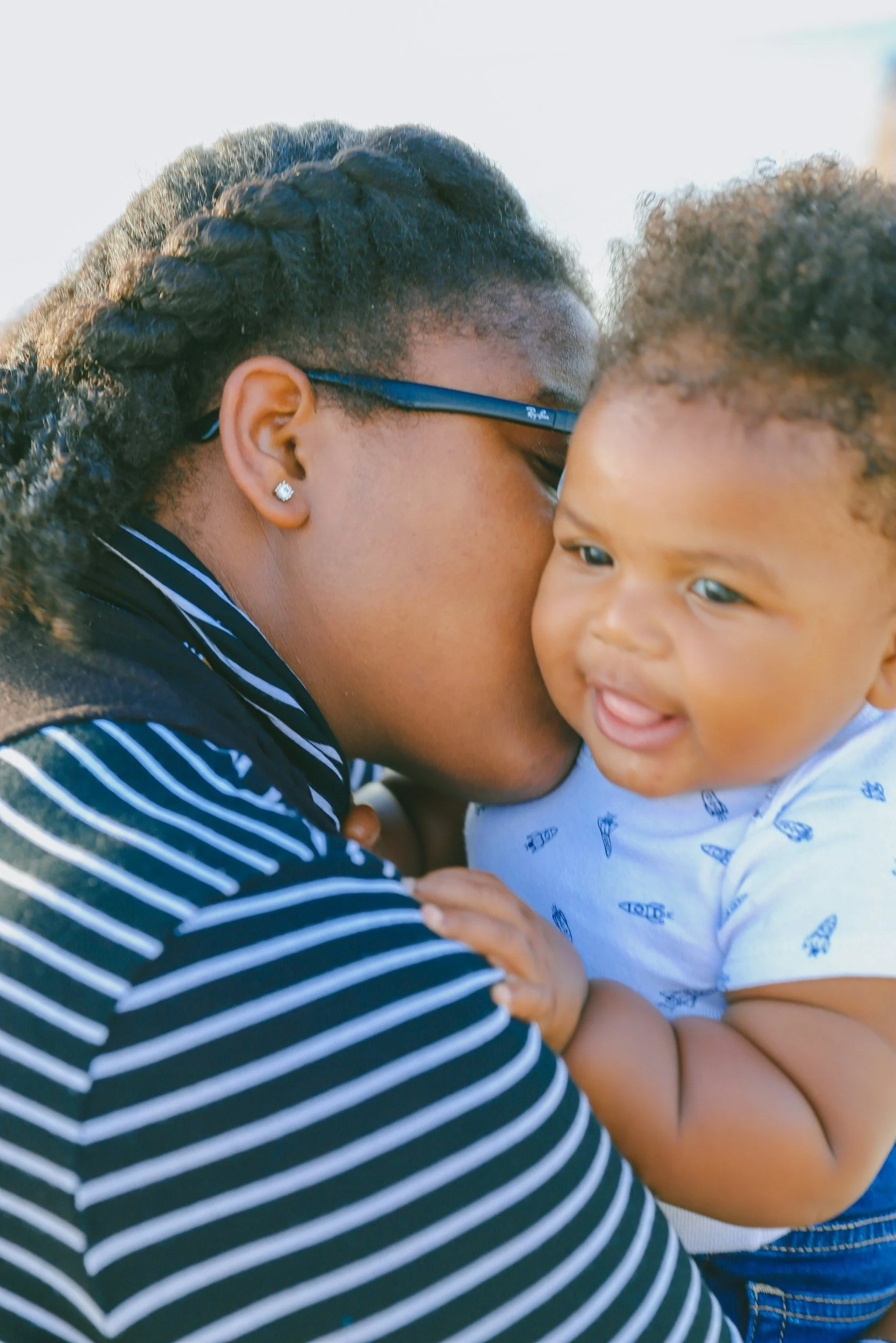 A woman with earrings and glasses kisses a young child on the cheek. The child is smiling slightly with their tongue out, wearing a white shirt with small blue patterns.