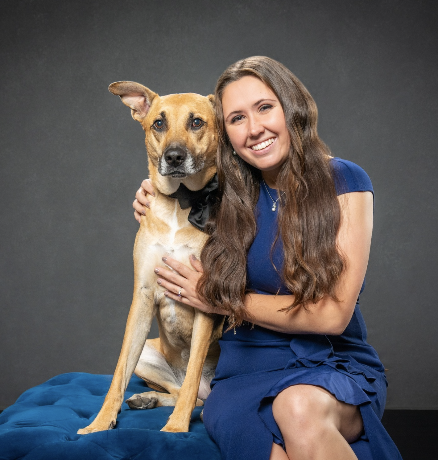 A smiling woman in a blue dress sitting on a blue blanket with a brown dog wearing a black bowtie, both looking at the camera against a plain dark background.