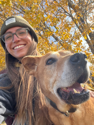 A woman smiling wearing a beanie hat and glasses, posing with a large, happy, brown dog outdoors during fall with yellow and orange leaves in the background.