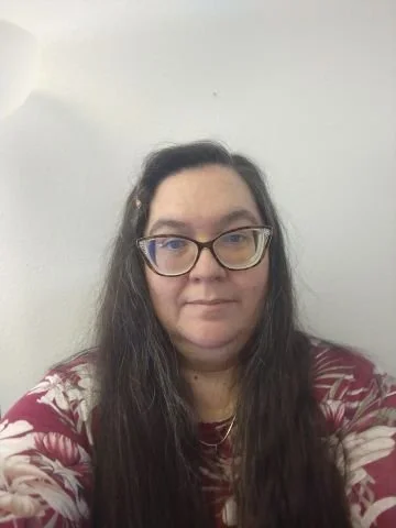 A woman with long dark hair wearing glasses and a red and white patterned shirt, standing against a plain white wall.