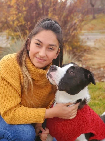 Young woman smiling and hugging a large mixed-breed dog with a bow tie, sitting on a chair outside a house.