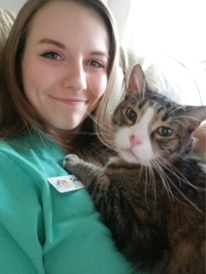 A woman smiling while holding a tabby cat with distinctive markings close to her face.