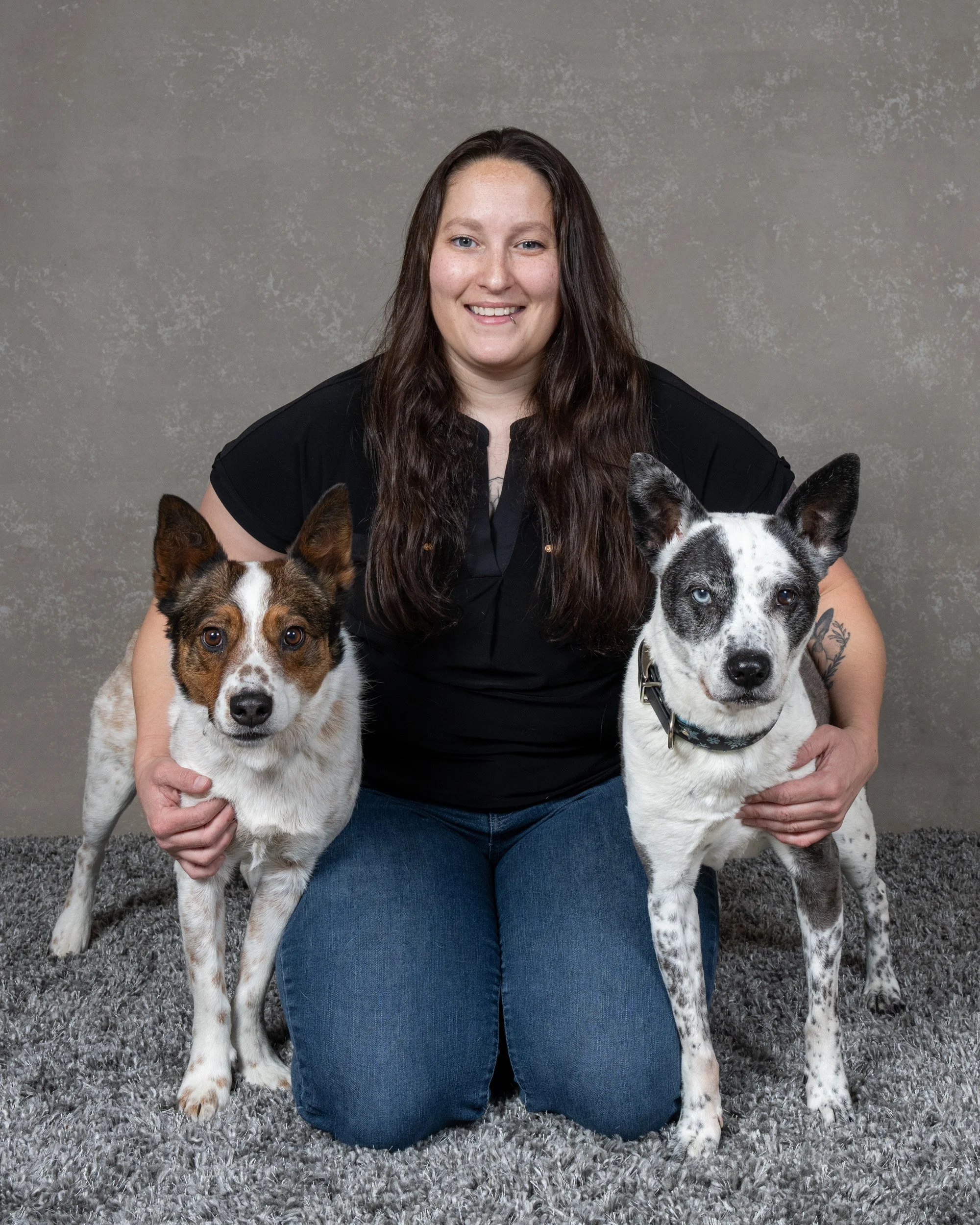 A woman with long brown hair kneeling on a gray carpet, holding two dogs on each side, with a neutral gray background.