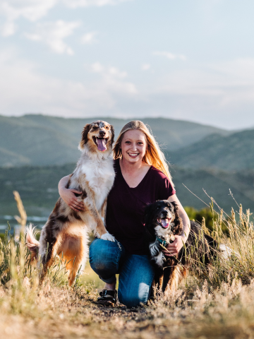 A woman smiling and kneeling outdoors in a grassy field with two dogs, one large and one small, scenic mountains in the background.