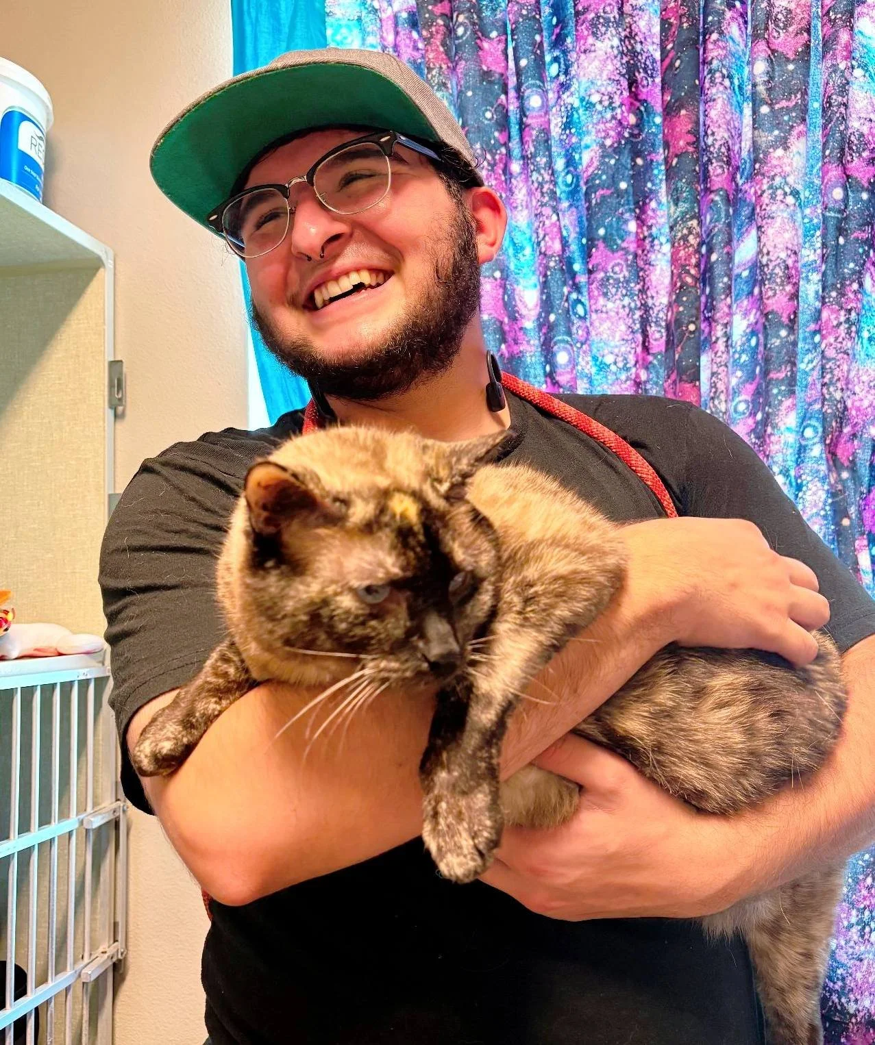 A man with glasses and a cap smiling while holding a calico cat in his arms.
