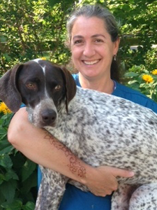 A woman smiling while holding a black and white dog outdoors in a garden with green foliage and yellow flowers.