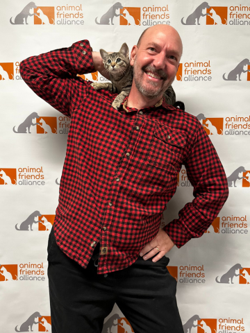 A man wearing a red and black checkered shirt smiling with a gray cat on his shoulder, standing in front of a backdrop with the Animal Friends Alliance logo.