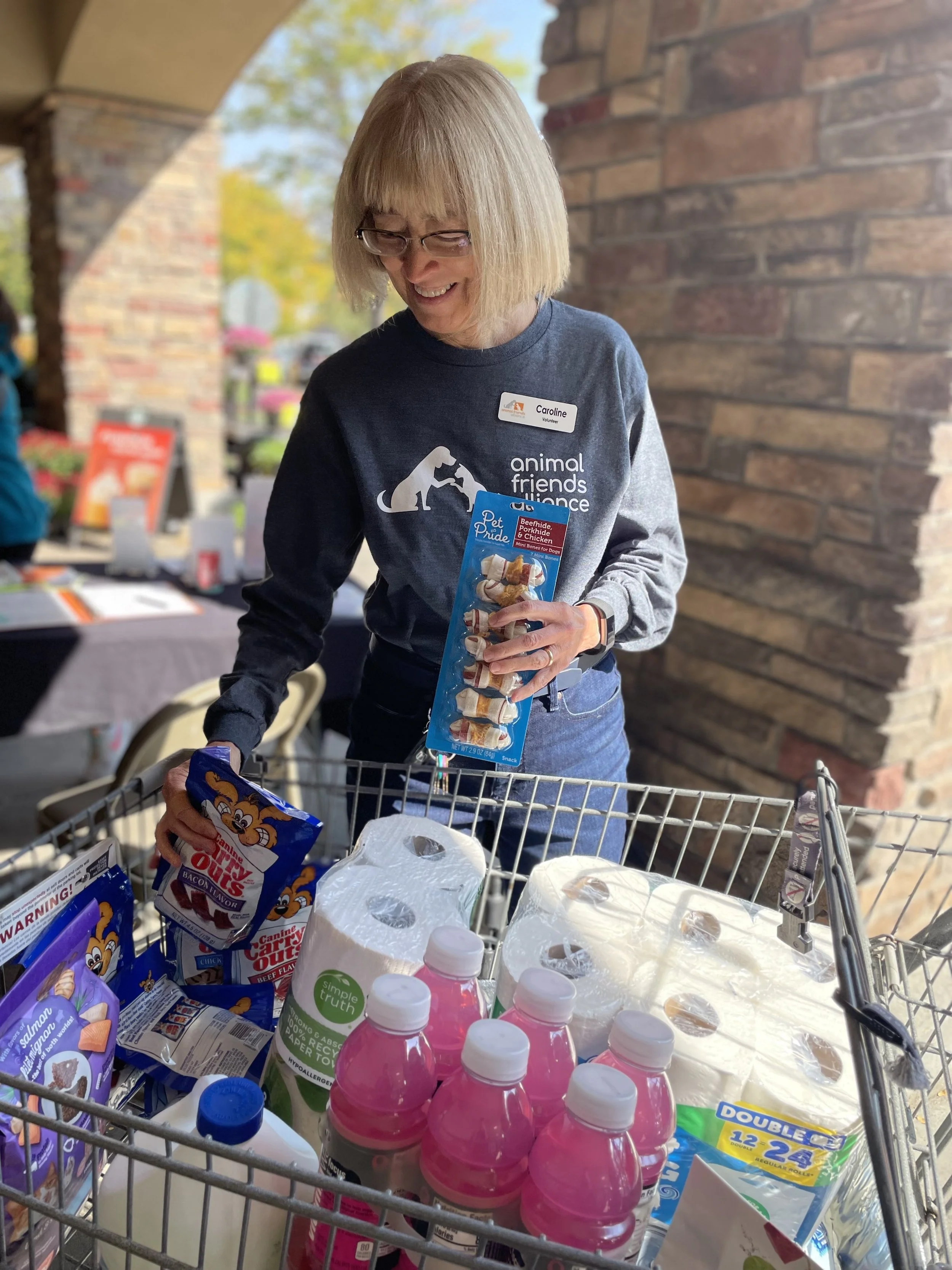 A smiling woman with glasses and a short blonde bob haircut shopping with a cart filled with paper towels, pink bottled drinks, and pet treats outside a store. She is wearing a gray sweatshirt with a dog logo and a name tag that says 'Caroline'.